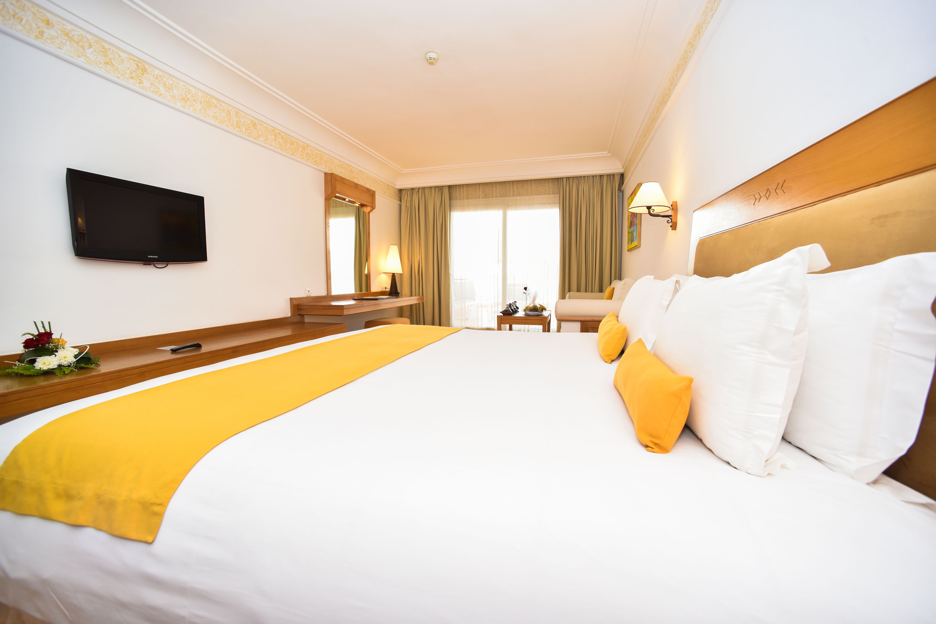 A hotel room with a large bed, a TV mounted on the wall, a desk with a lamp, and a chair.