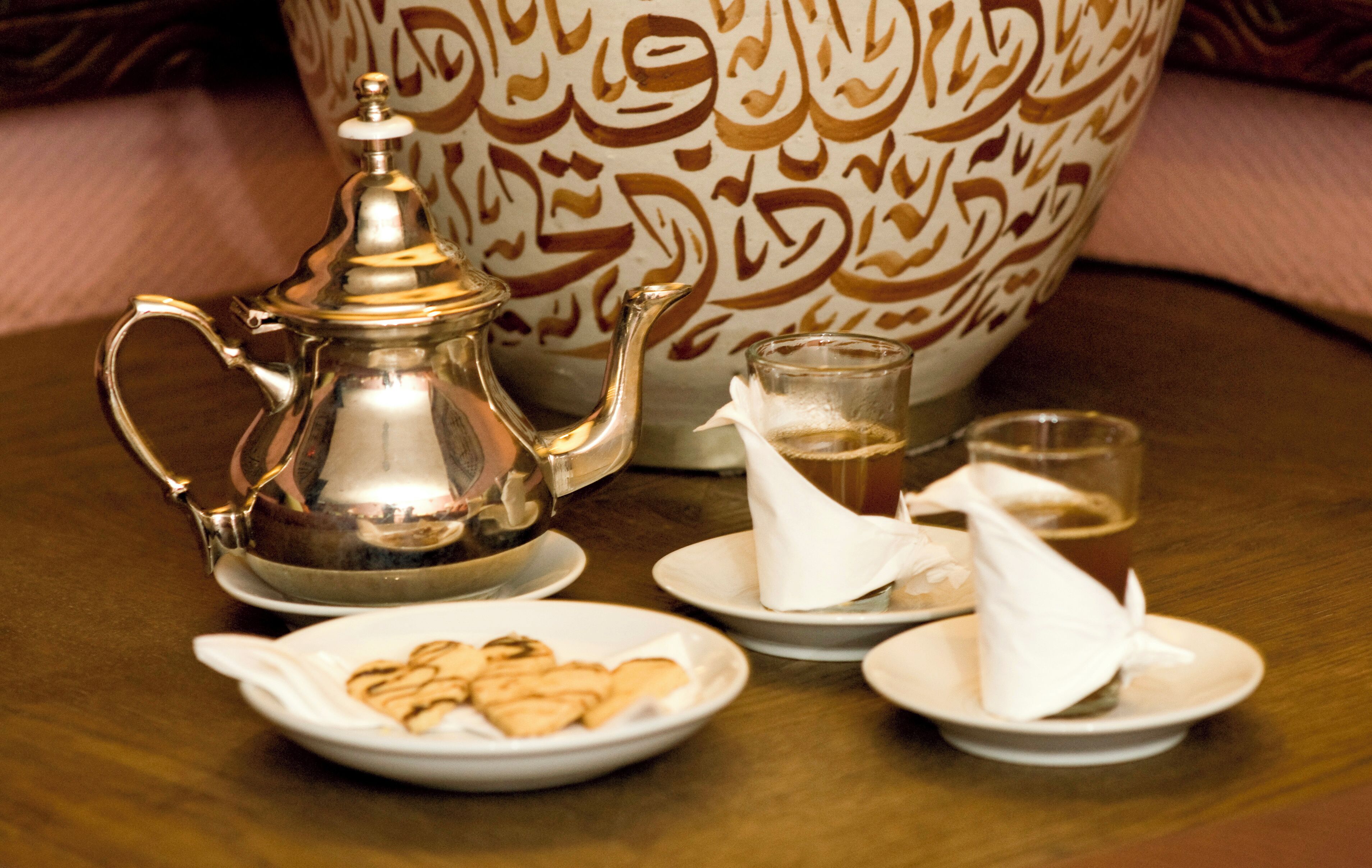 A traditional tea set with a silver teapot and cups, accompanied by plates of cookies and a decorative bowl in the background.