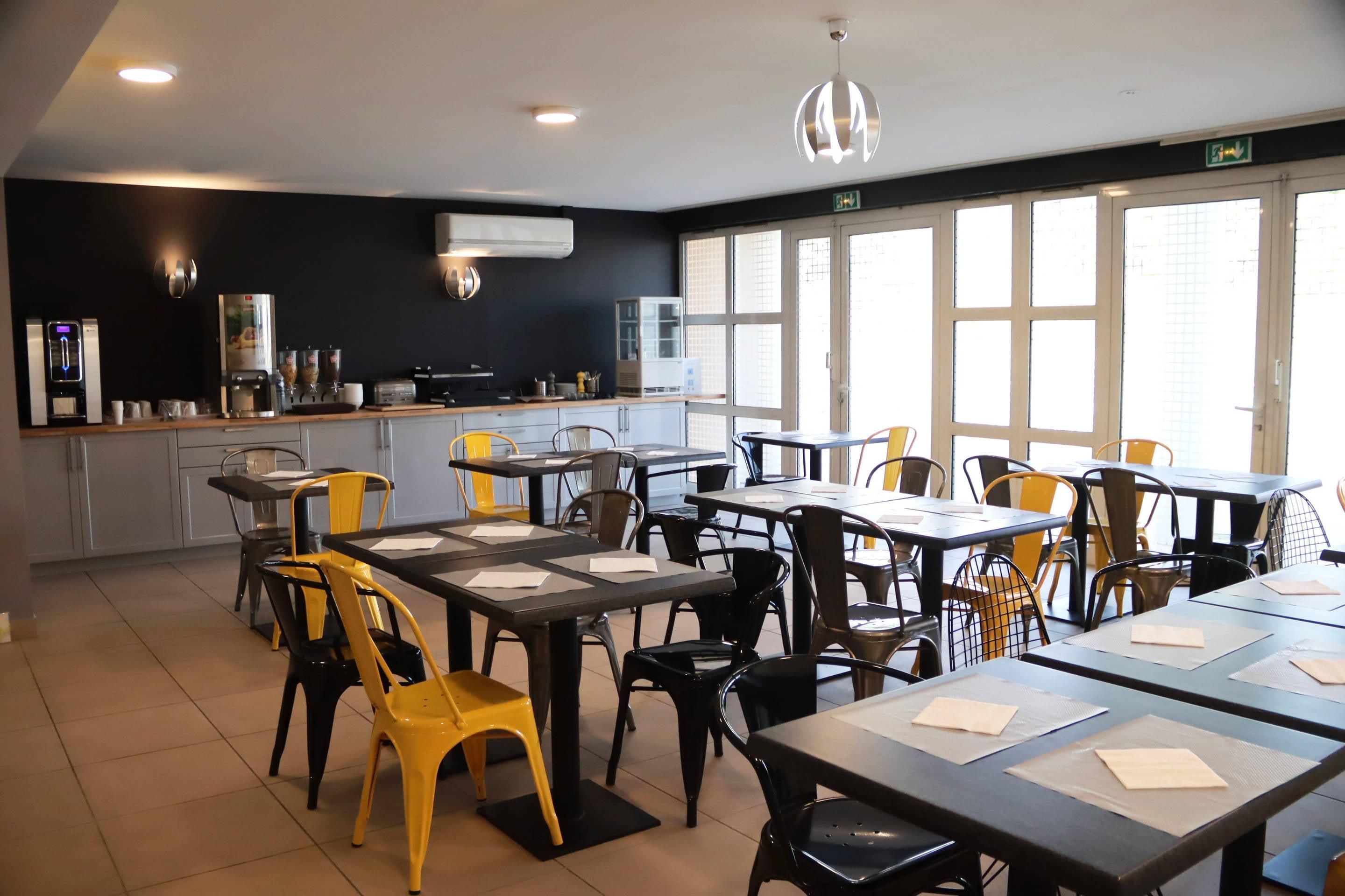 Comedor con mesas y sillas, zona de cocina con electrodomésticos y ventanas grandes.