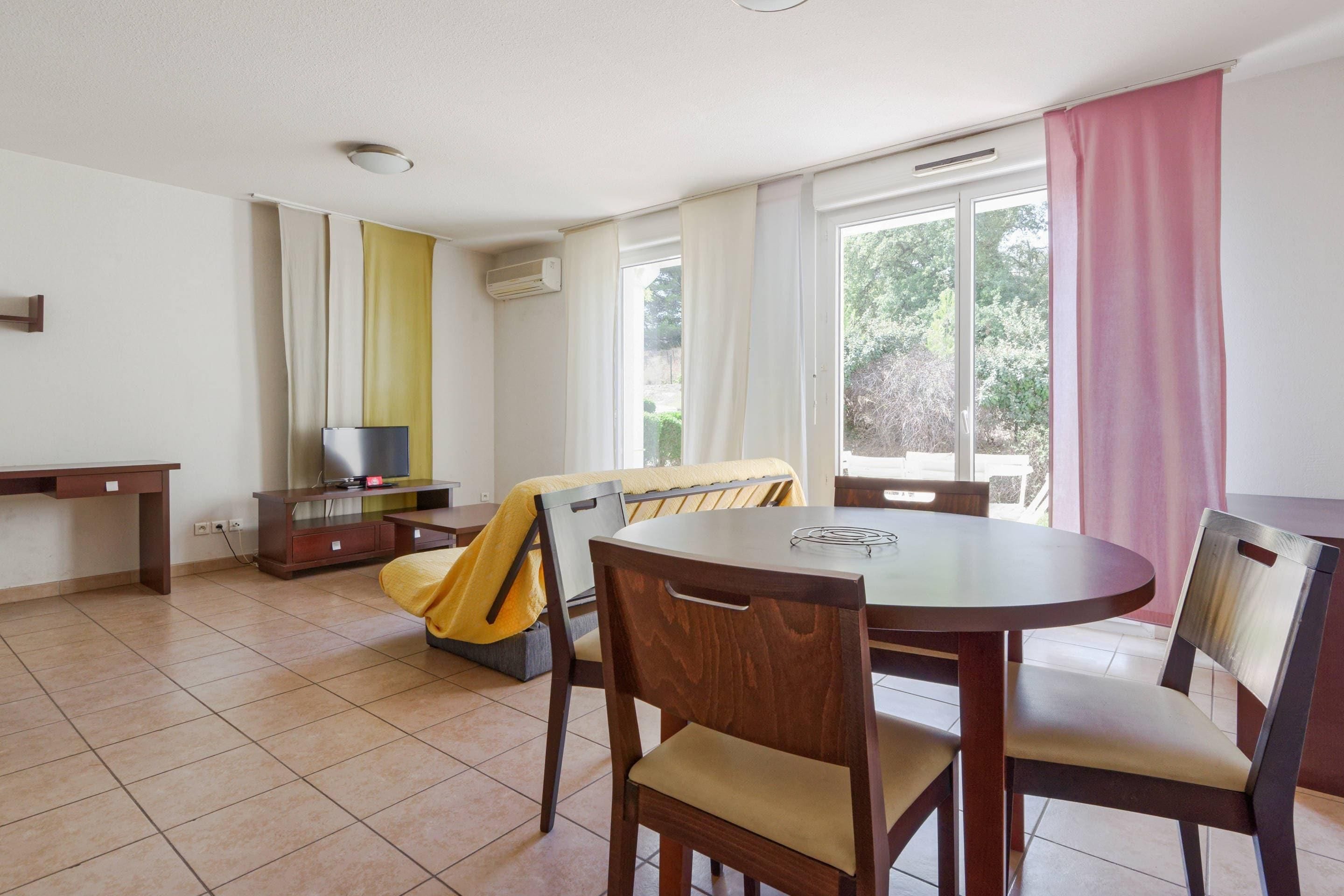Ein Zimmer mit rundem Tisch, zwei Stühlen, einer Couch, einem Fernseher und einem Fenster mit Vorhängen.