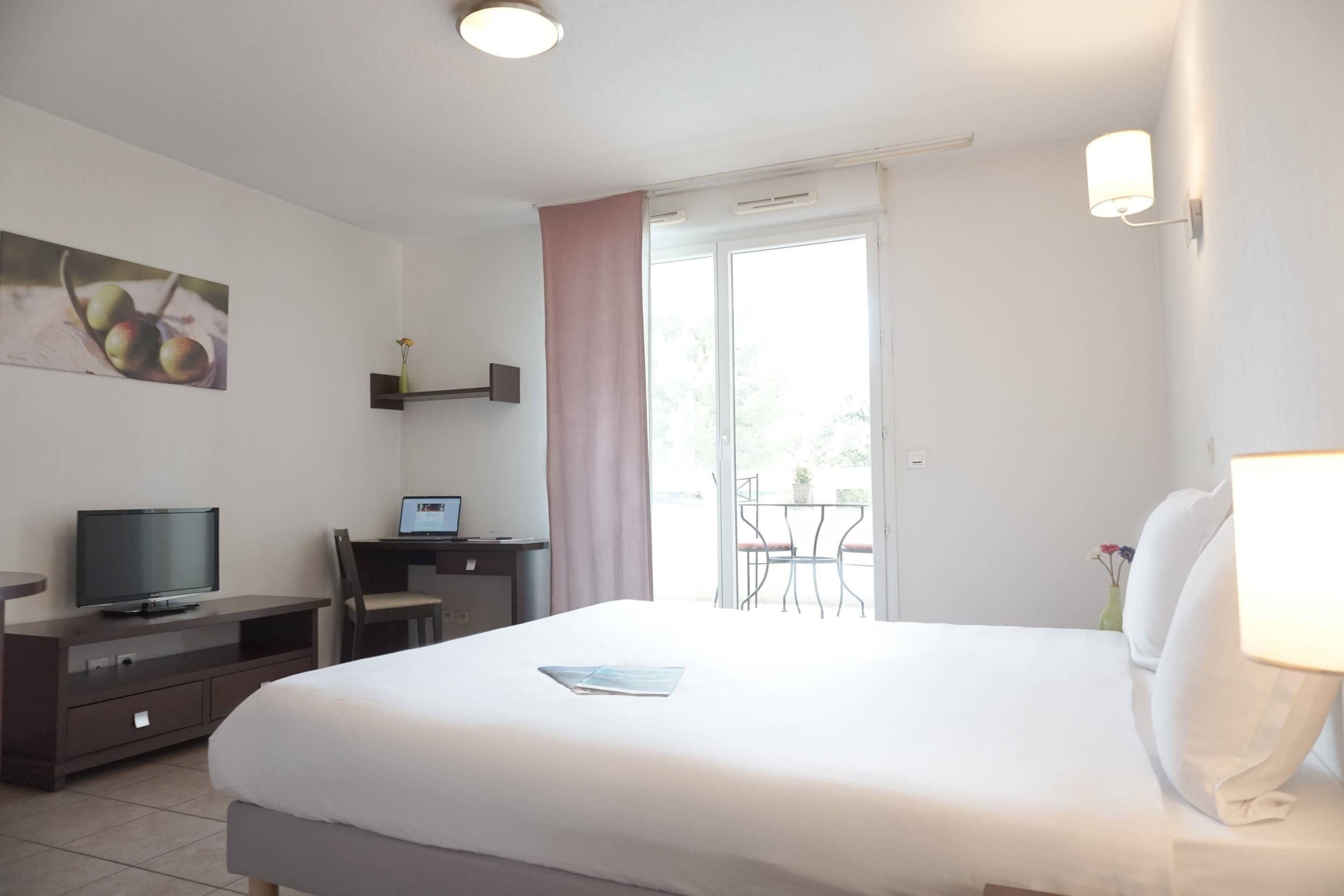 A hotel room with a bed, a desk with a computer, a television, and a balcony with a table and chairs.
