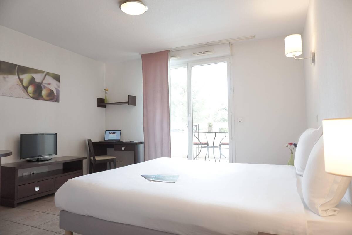 A hotel room with a bed, a desk with a computer, a television, and a balcony with a table and chairs.