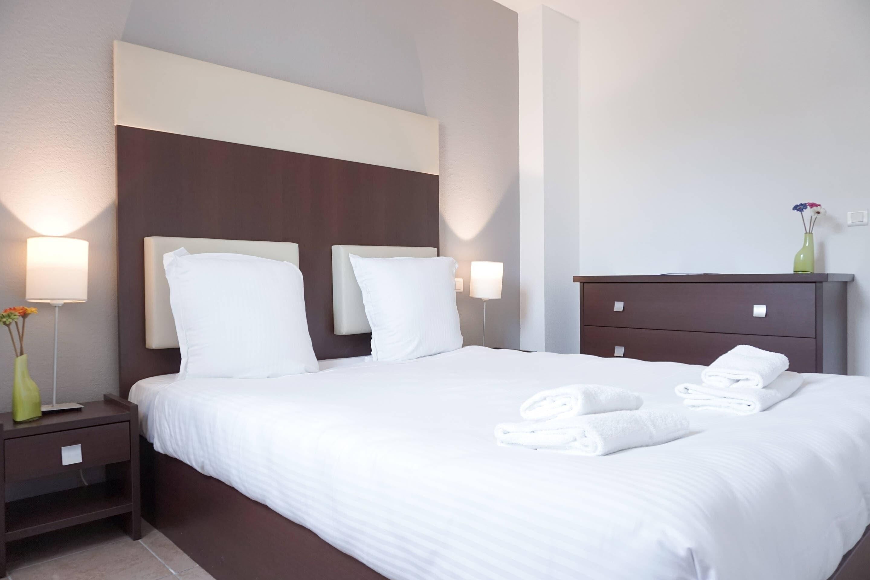 A hotel room with a large bed, two bedside lamps, a nightstand, a dresser, and a vase with flowers.