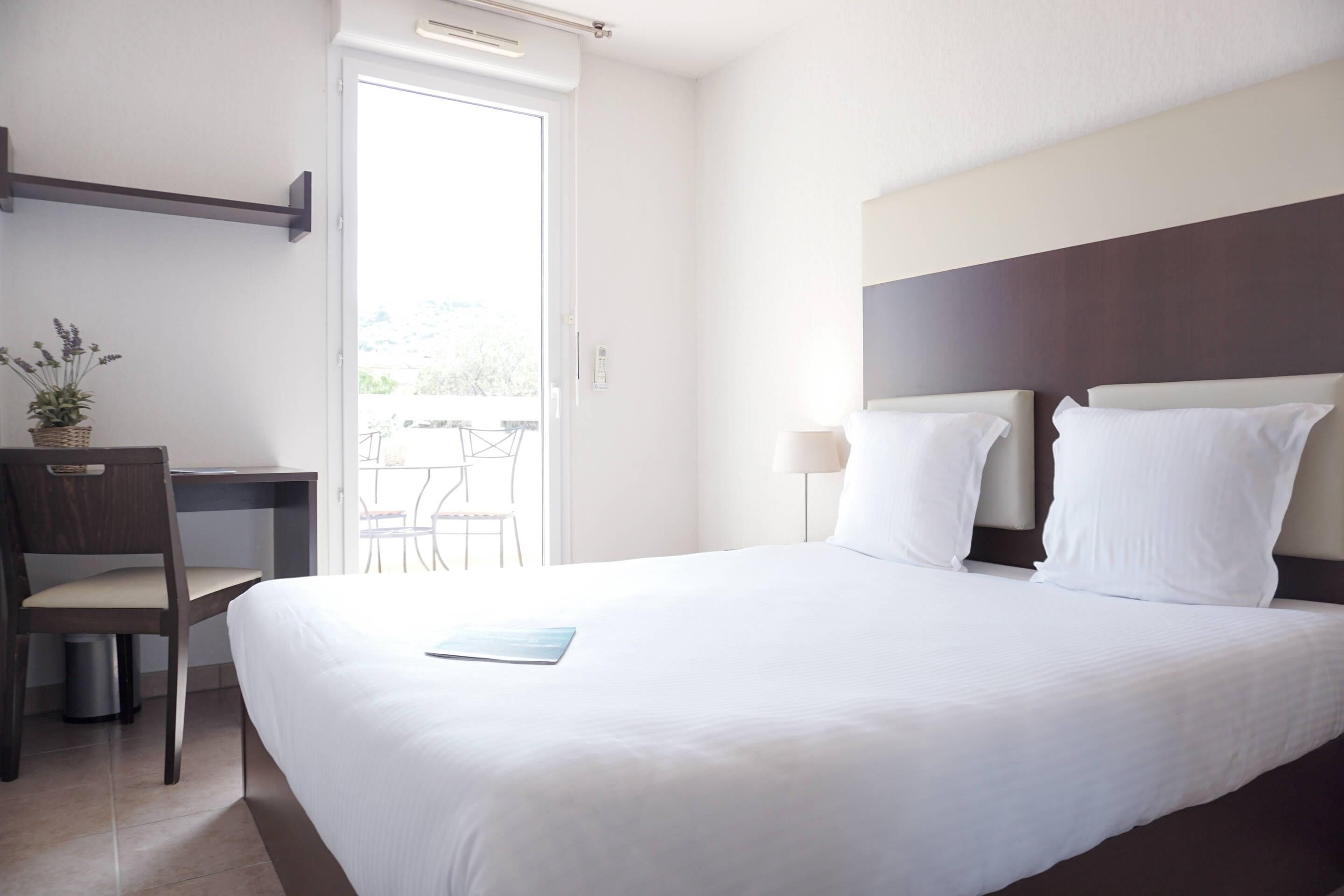 A hotel room with a bed, a desk, a chair, a small potted plant, and a view of the outdoors.