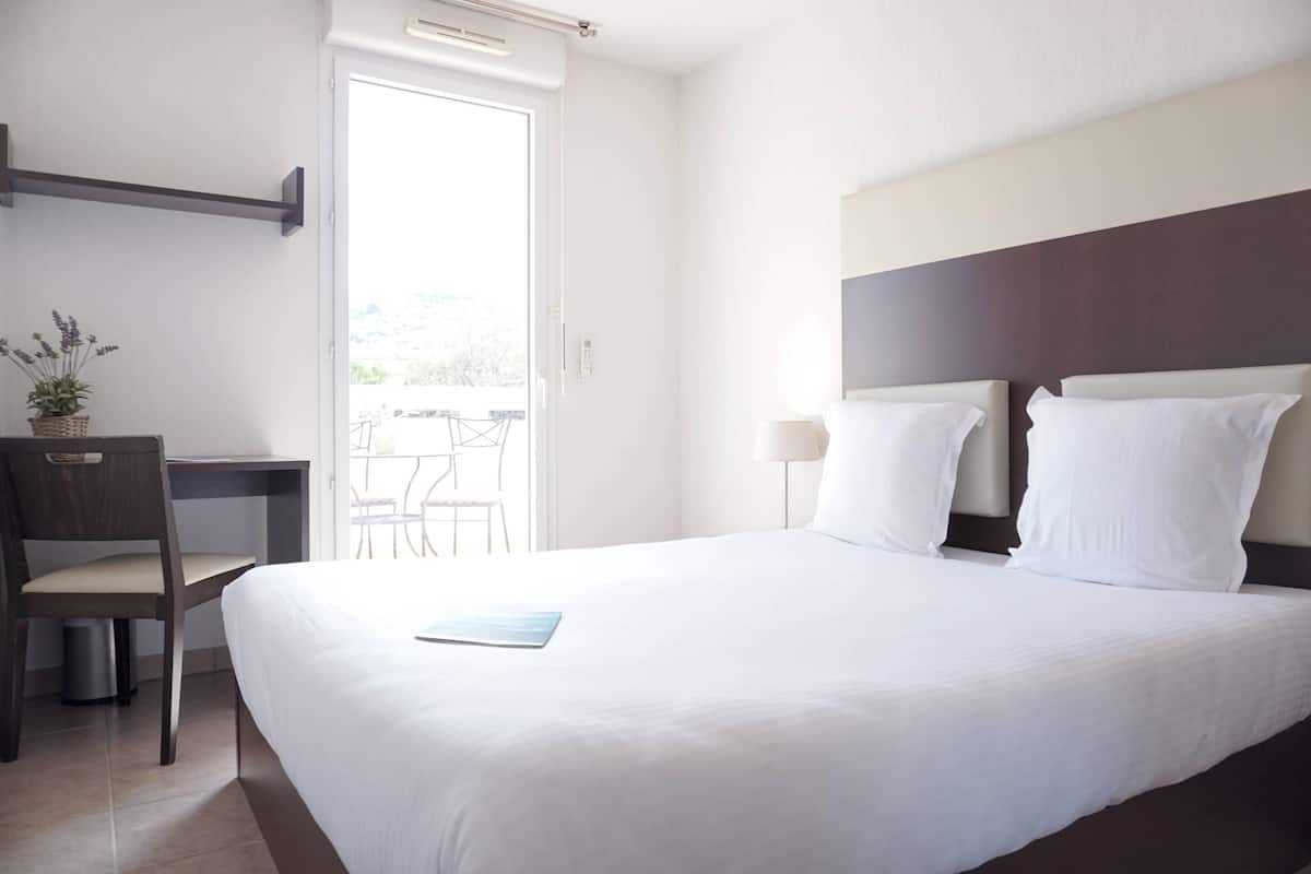 A hotel room with a bed, a desk, a chair, a small potted plant, and a view of the outdoors.