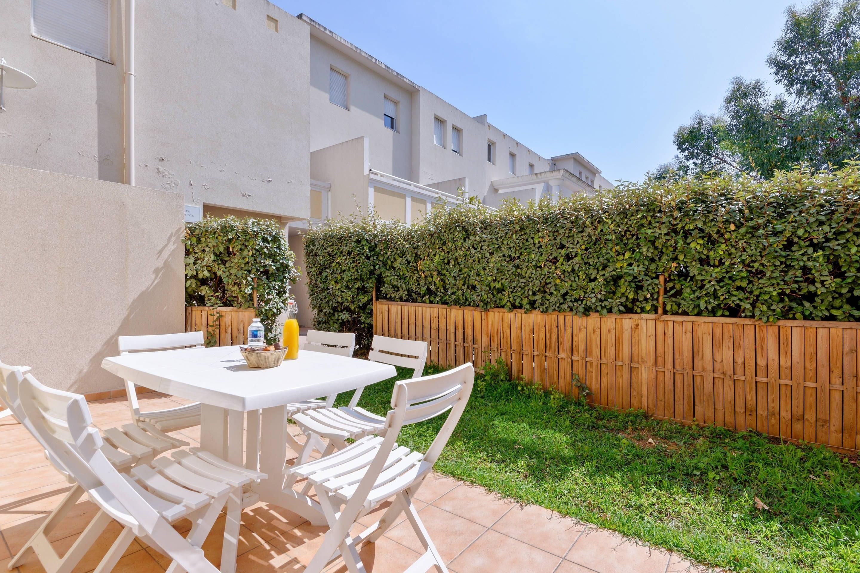 A patio with a white table and chairs, a wooden fence, and a hedge.