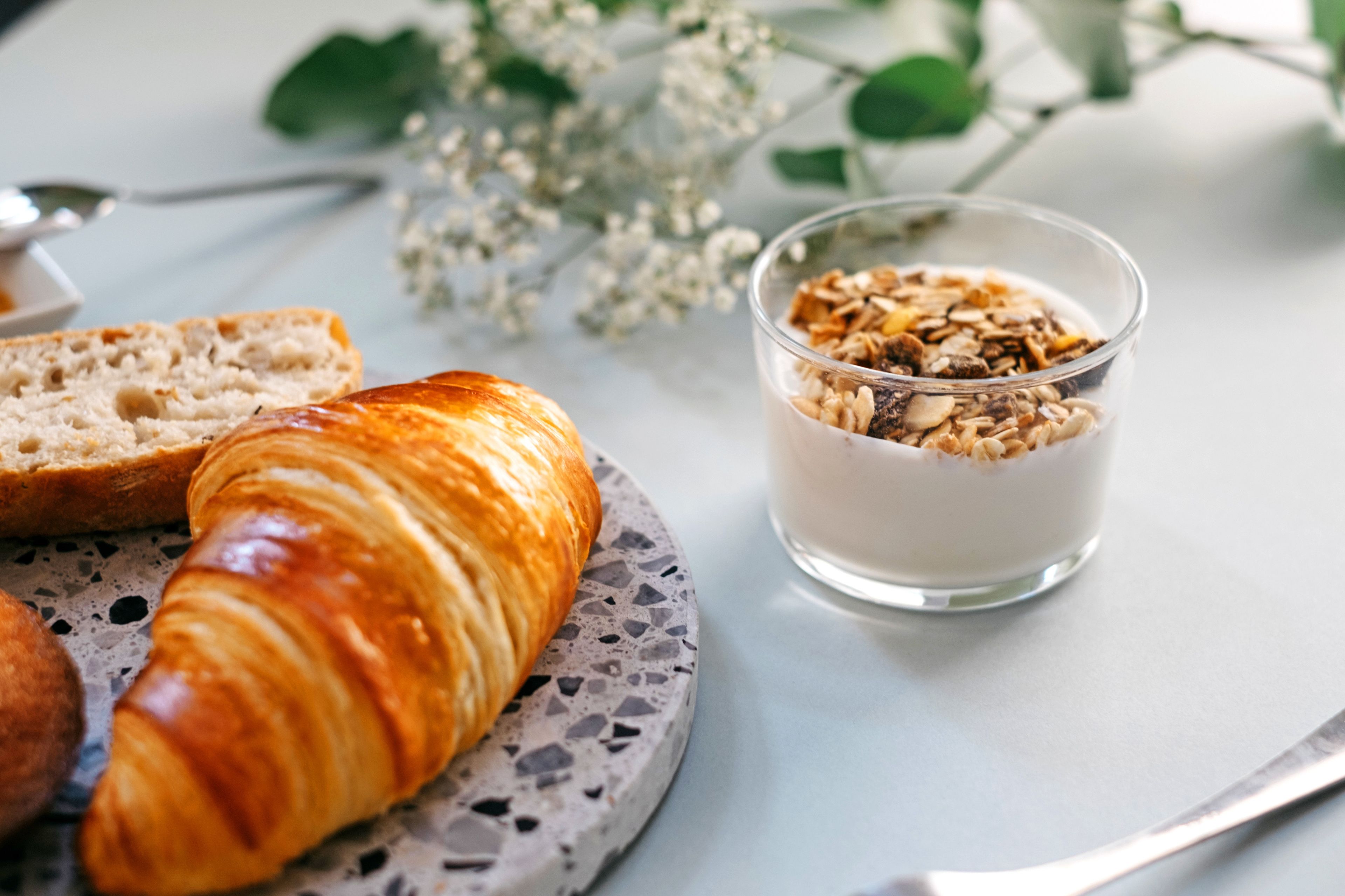 Een croissant, een sneetje brood en een glas yoghurt met granola op een tafel.