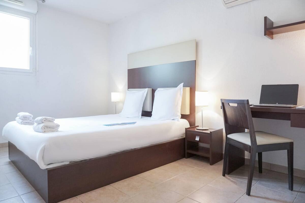 A hotel room with a bed, a desk, a chair, and a laptop.