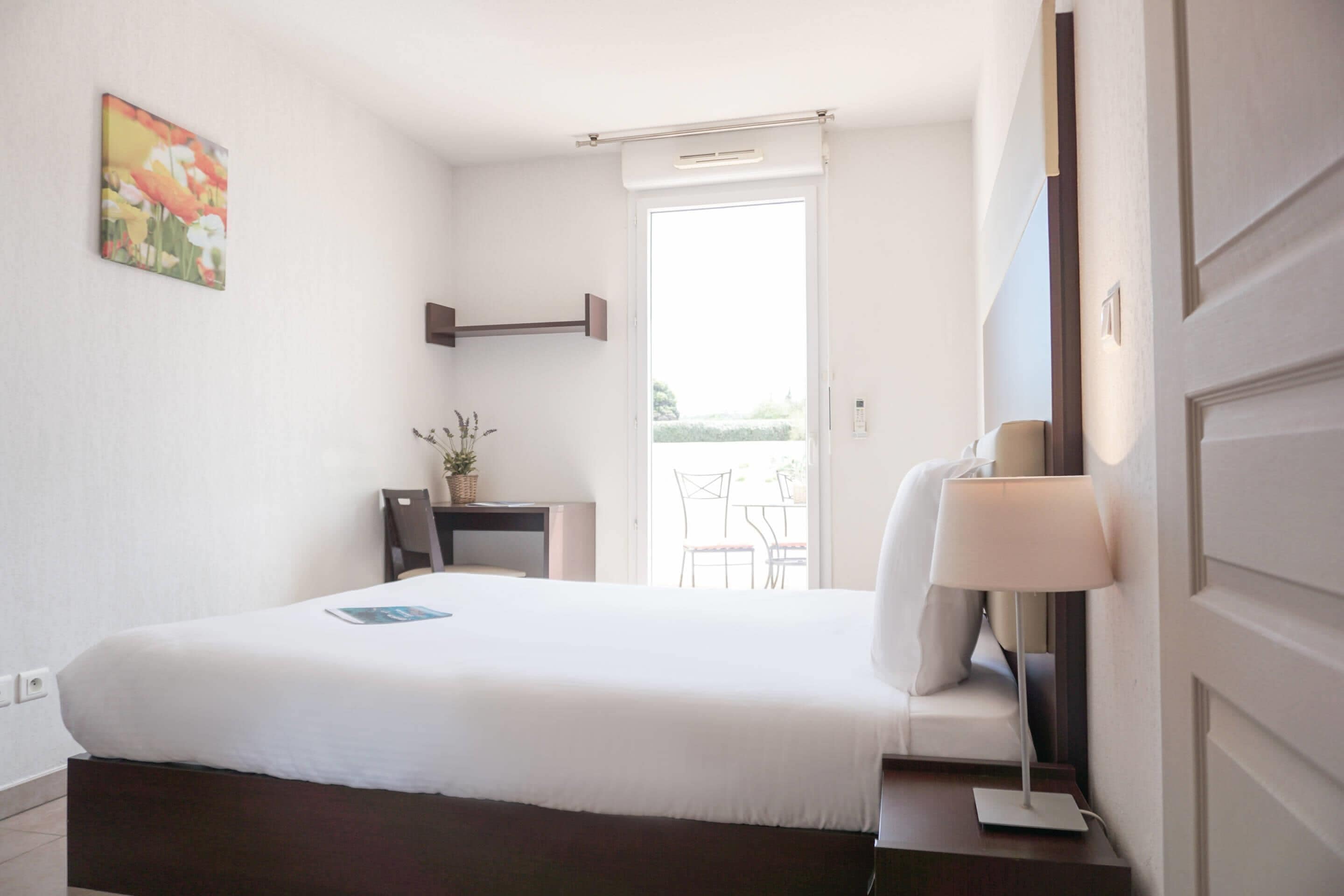 A hotel room with a bed, a desk, a chair, a lamp, and a window with a view of the outdoors.