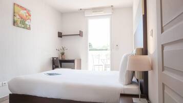 A hotel room with a bed, a desk, a chair, a lamp, and a window with a view of the outdoors.