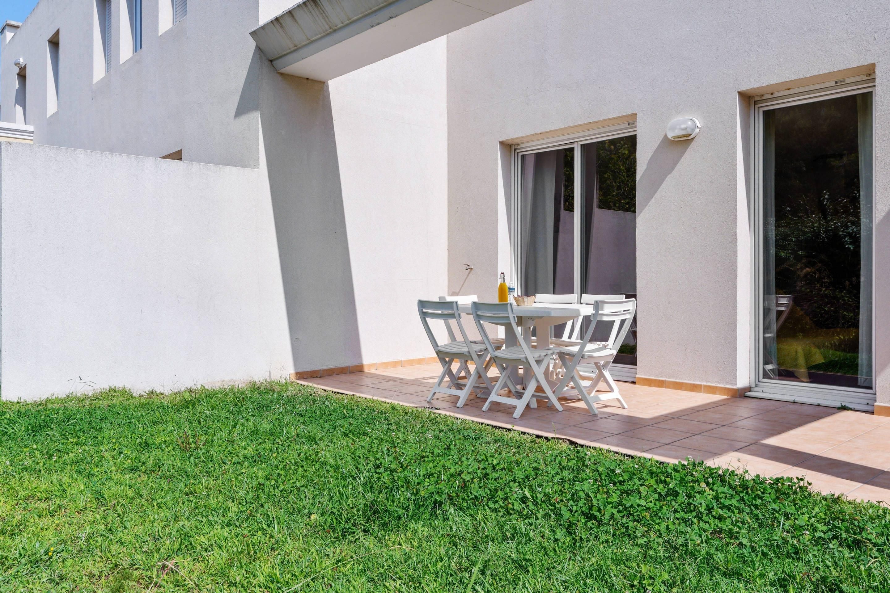 A modern outdoor patio with a table and chairs, a small garden, and a building with large windows and a sliding door.