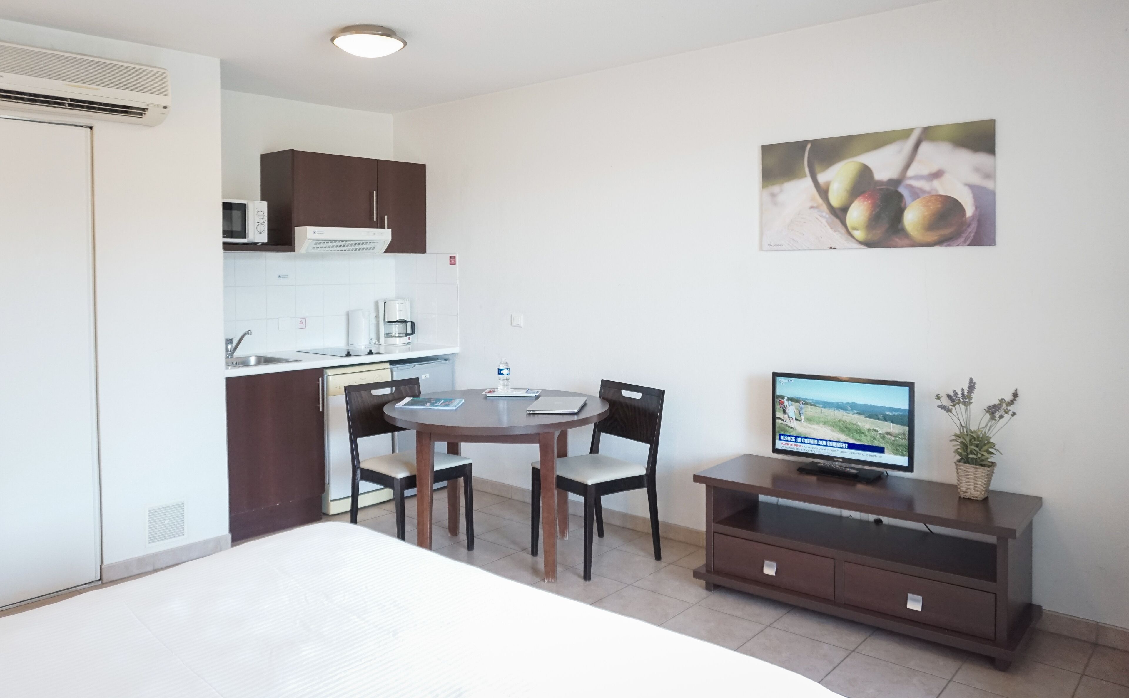 A compact hotel room with a bed, a dining table with chairs, a kitchenette, a television, and a small cabinet.