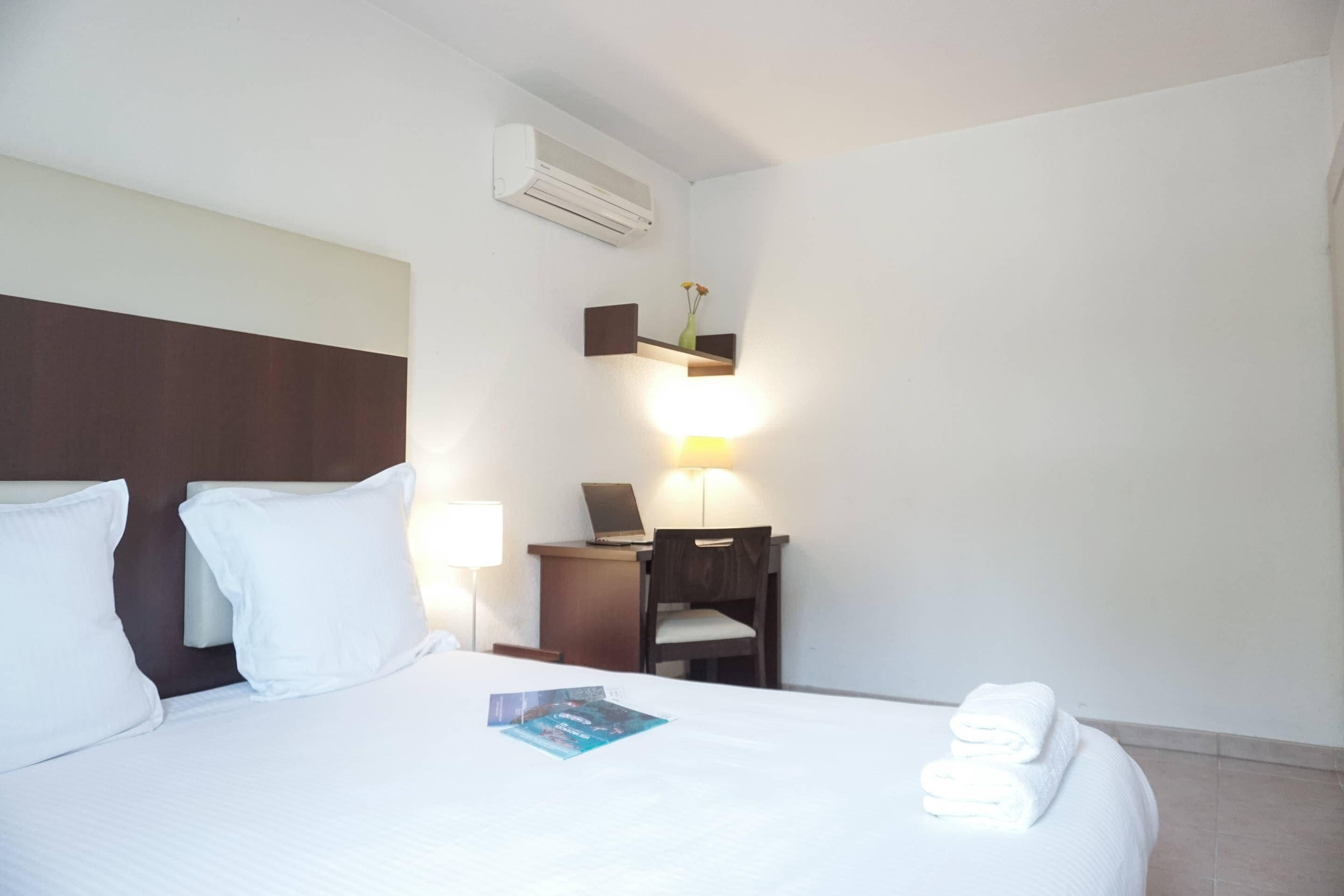 A hotel room with a bed, pillows, a desk with a laptop, a chair, a lamp, and a wall-mounted shelf.