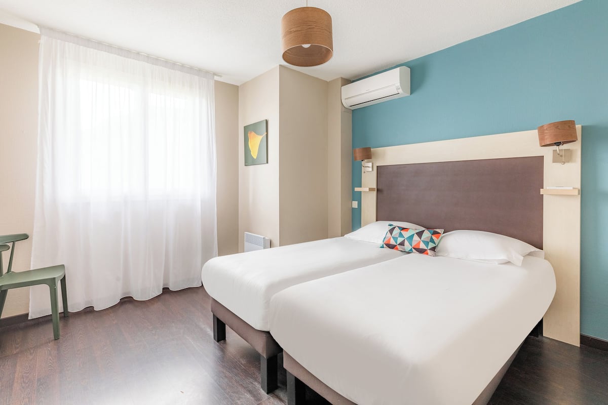 Ein Hotelzimmer mit zwei Betten, einem Schreibtisch, einem Stuhl, einem Fenster mit durchsichtigen Vorhängen, einer Klimaanlage und einer Wandlampe.