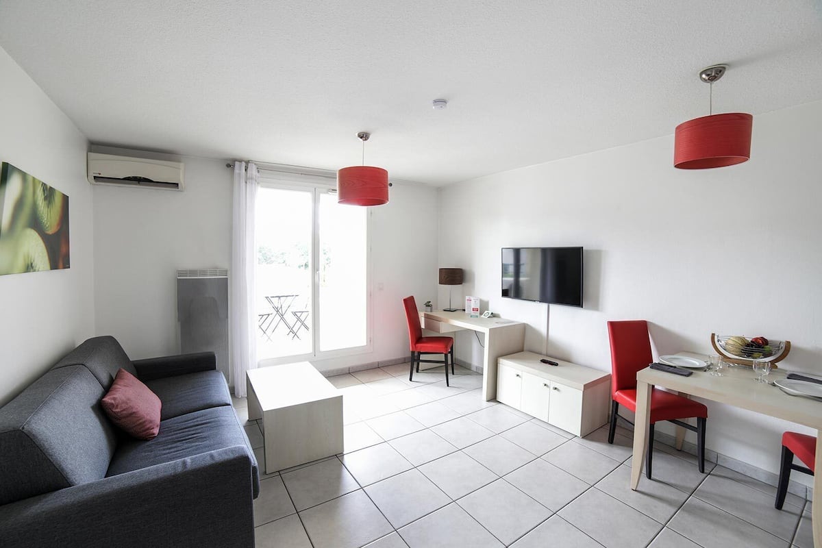 Ein modernes, helles Wohnzimmer mit grauem Sofa, roten Sesseln, einem weißen Schreibtisch und einem Fernseher.