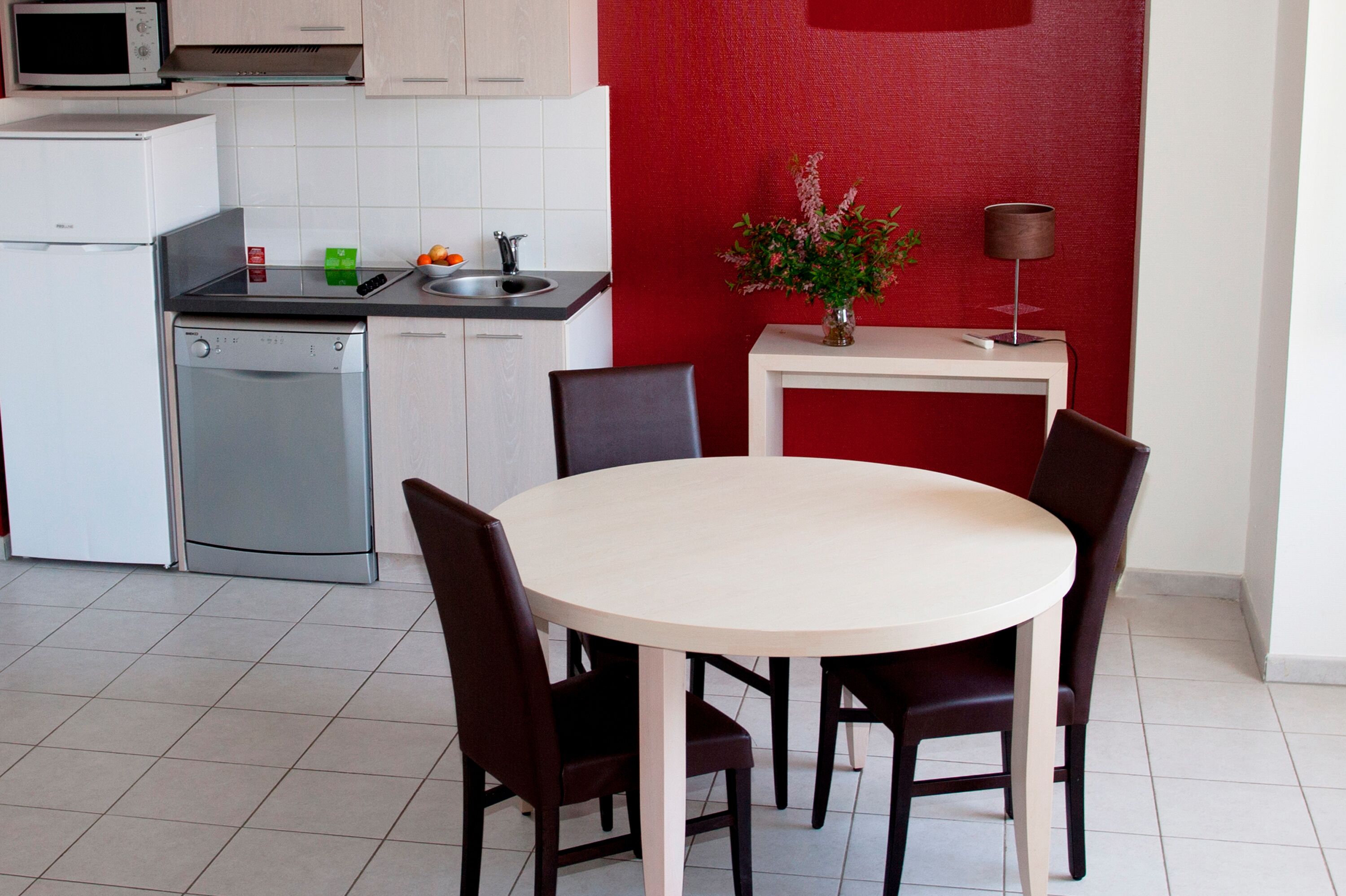 Una cocina compacta con microondas, pileta y lavavajillas. Una mesa de comedor con cuatro sillas se ubica frente a una pared roja decorativa.