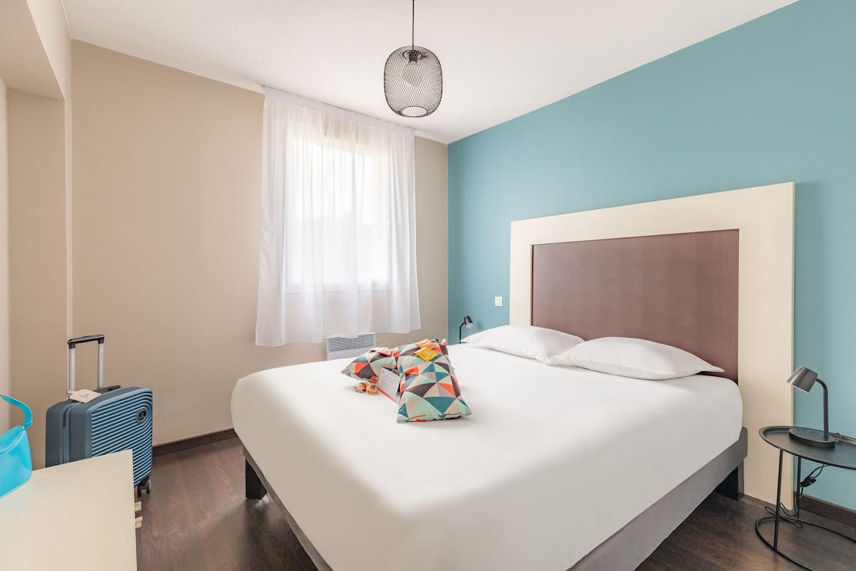 Ein Hotelzimmer mit einem großen Bett, einem Koffer, einem Schreibtisch und einem Fenster mit Vorhängen.