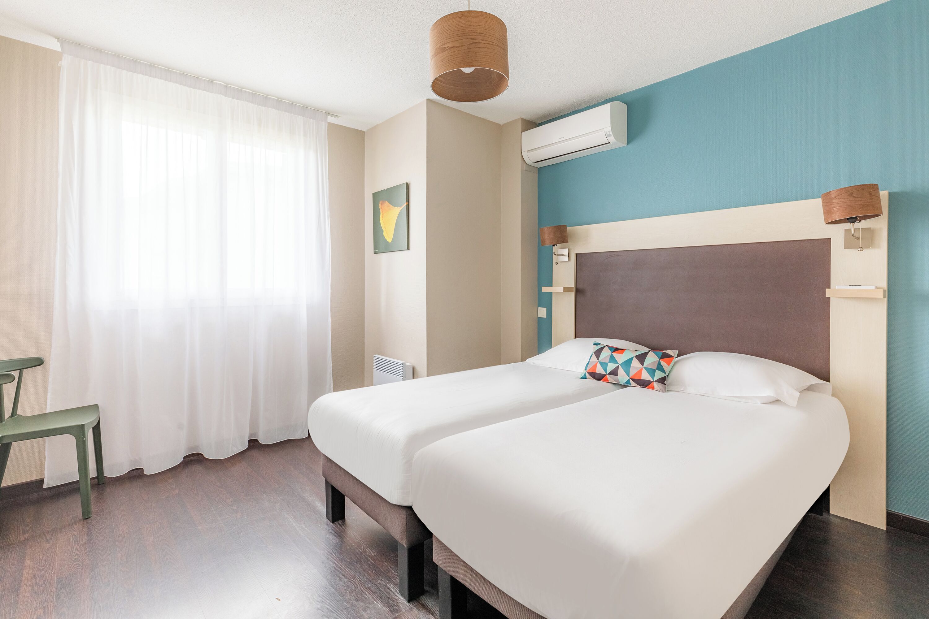 Une chambre d’hôtel avec deux lits, un bureau, une chaise, une fenêtre avec des rideaux, un climatiseur et une lampe fixée au mur.