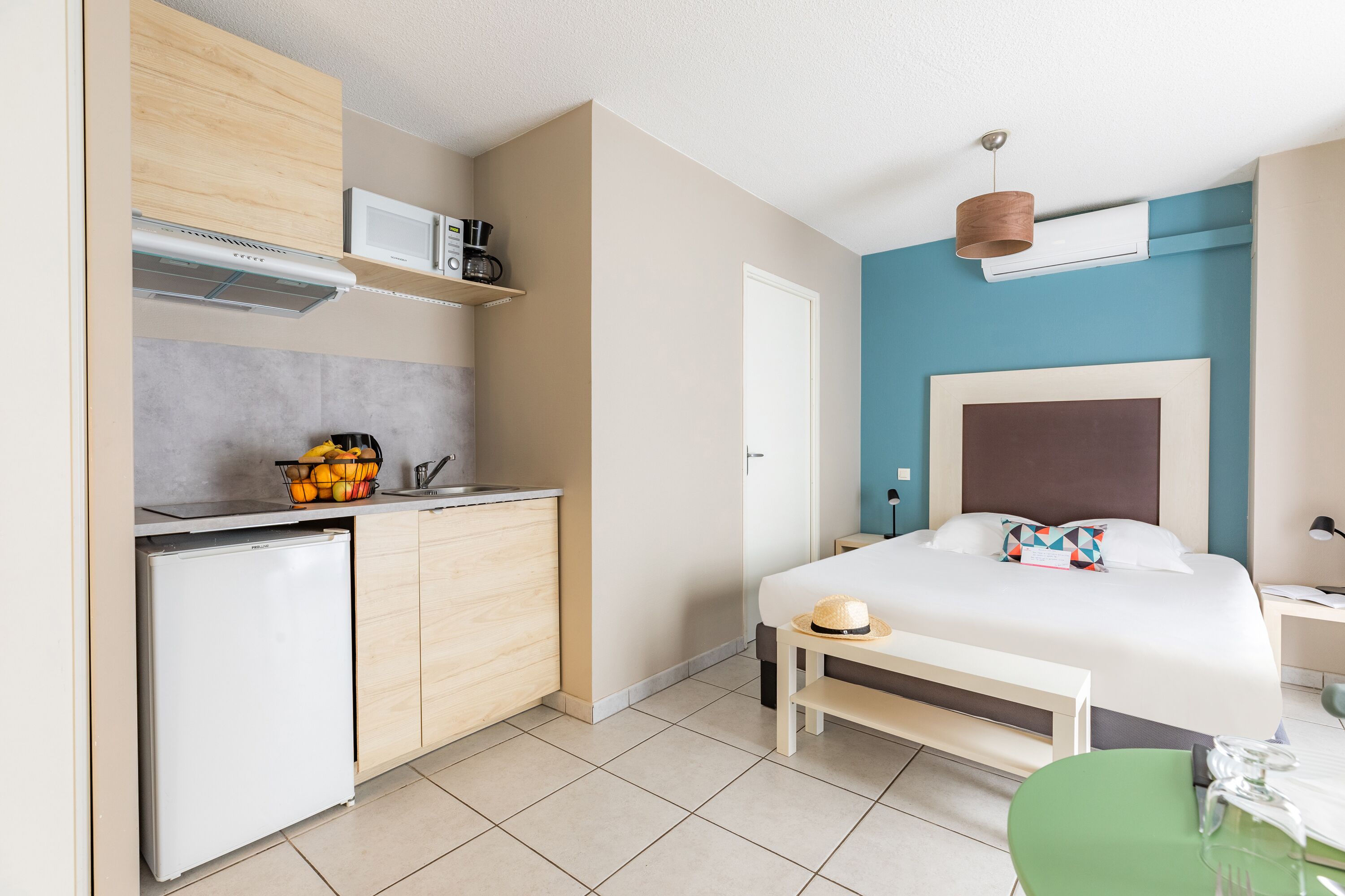 Een kleine hotelkamer met een kitchenette, een bed, nachtkastje en een zithoekje.