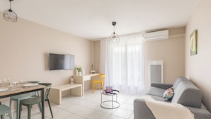 A modern living room with a grey sofa, a dining table with chairs, a small round table with a lamp, and a TV mounted on the wall. - Appart'City Classic Bordeaux Aéroport St Jean D'Illac (Saint-Jean-d'Illac)