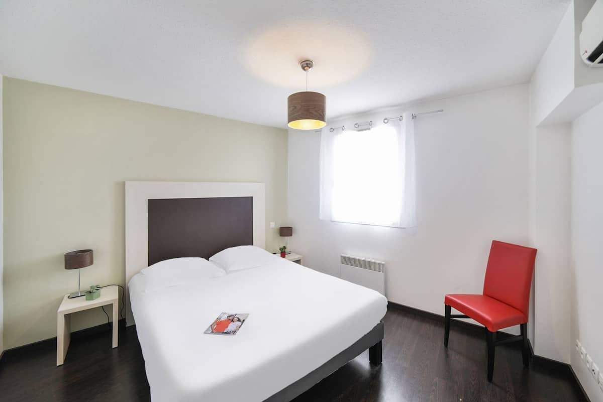 Ein Hotelzimmer mit Bett, Sessel, Nachttisch, Lampe, Fenster und Heizkörper.