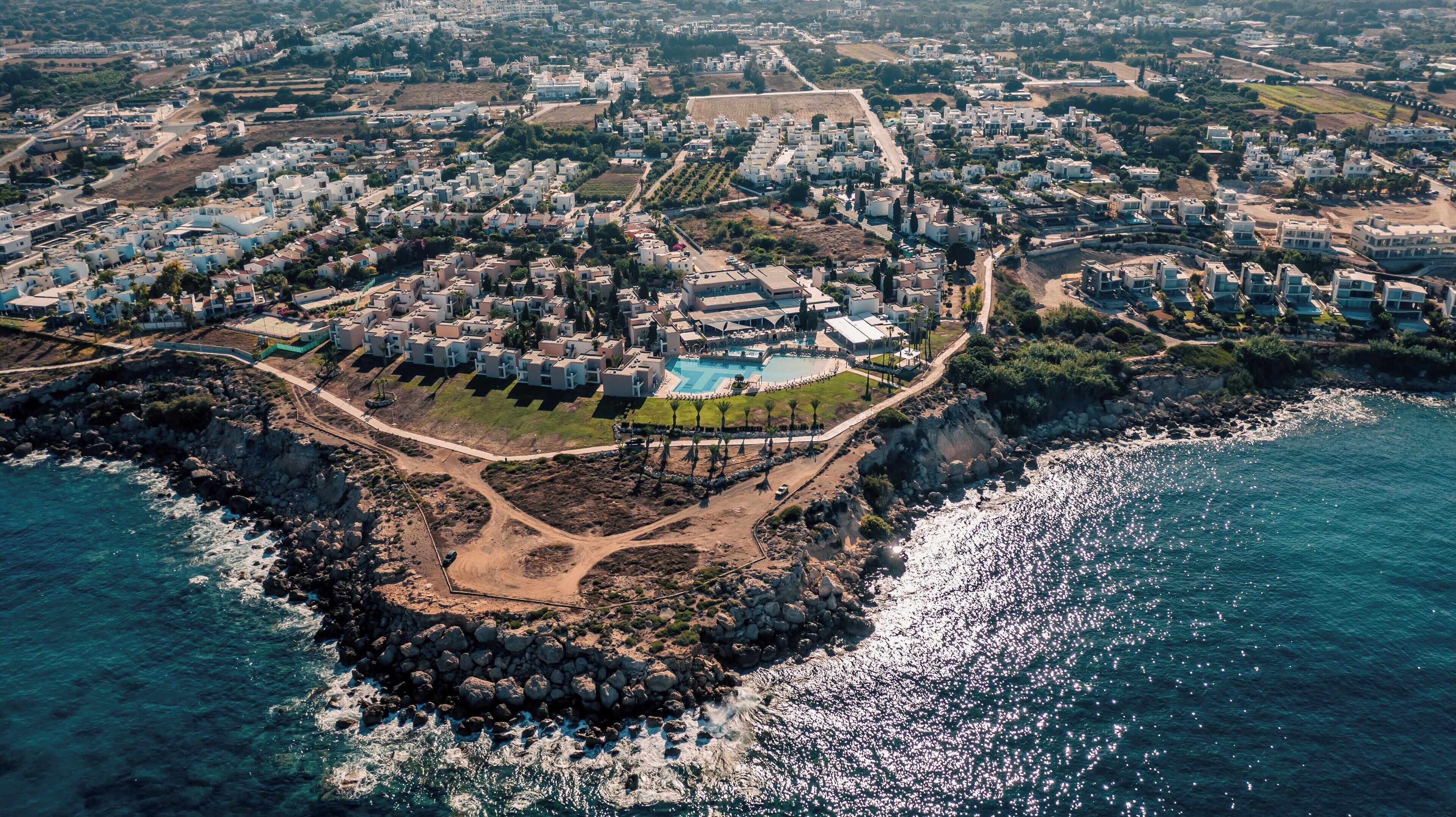 An aerial view of a coastal resort with multiple buildings, a central pool area, and rocky coastline.