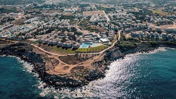 An aerial view of a coastal resort with multiple buildings, a central pool area, and rocky coastline.