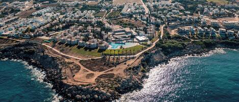An aerial view of a coastal resort with multiple buildings, a central pool area, and rocky coastline.