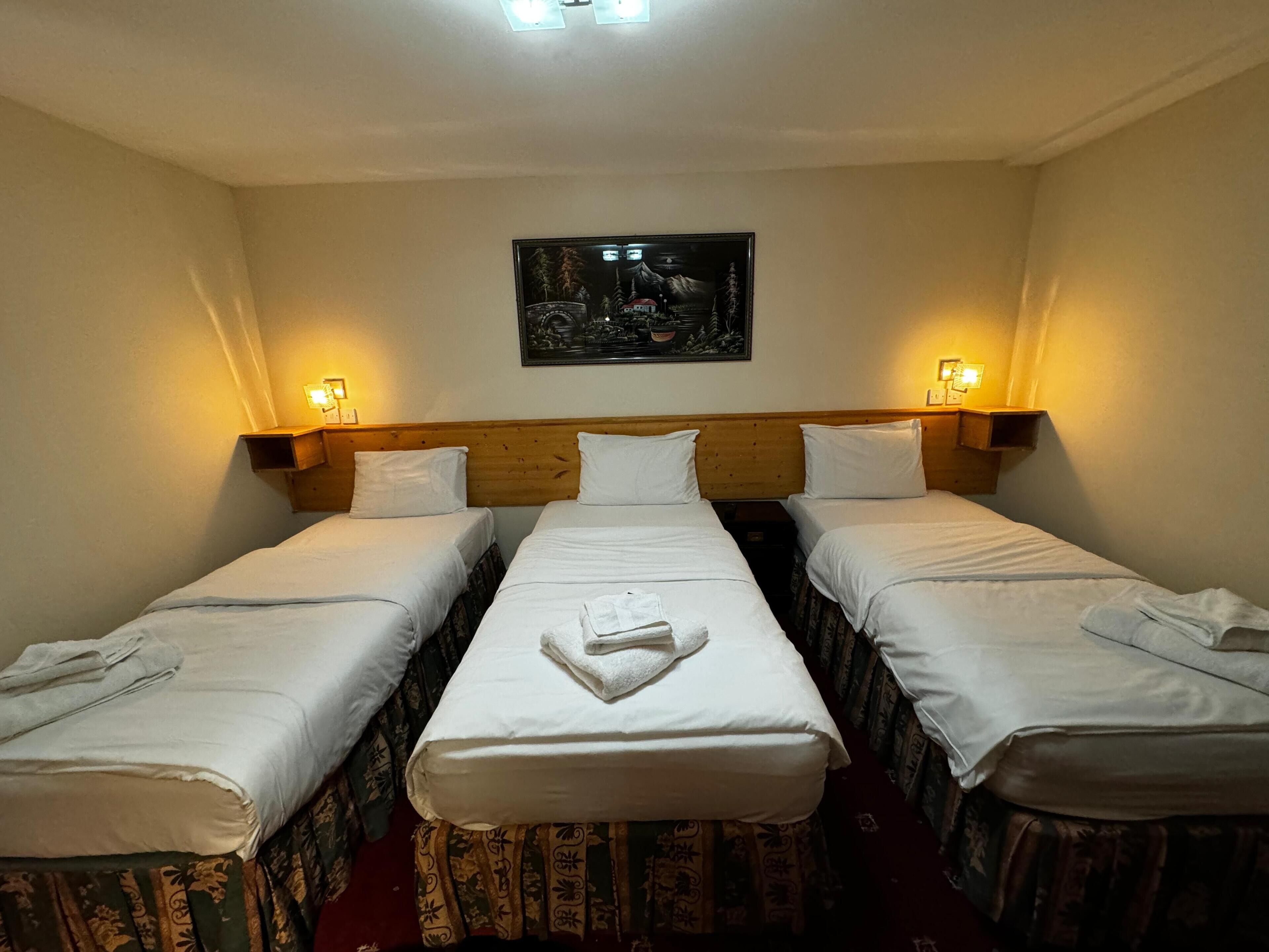 Quarto de hotel com duas camas de solteiro, cabeceira de madeira, quadro pendurado na parede e luminárias de cabeceira.