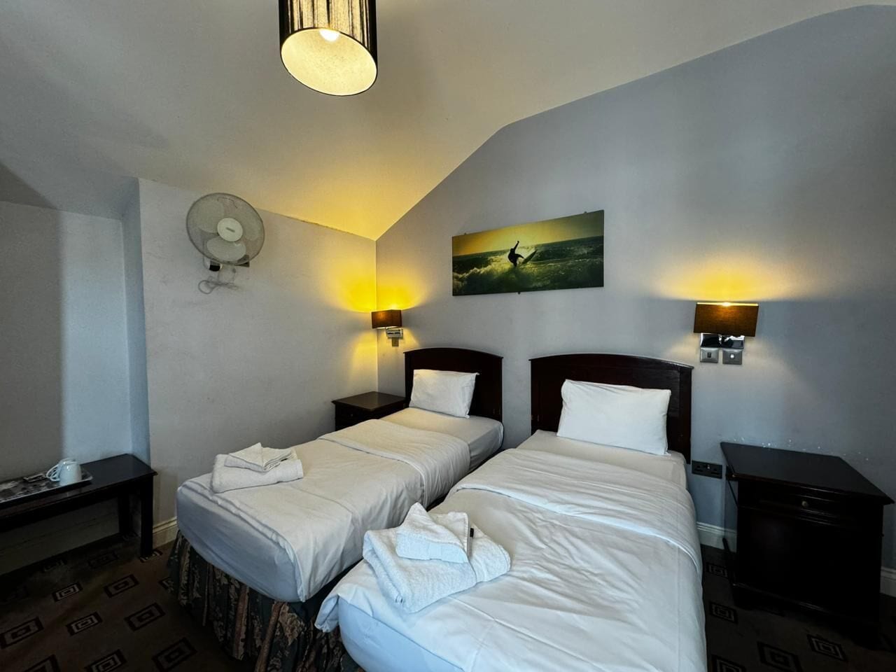 Een hotelkamer met twee bedden, een schilderij van een surfer en een ventilator aan de muur.