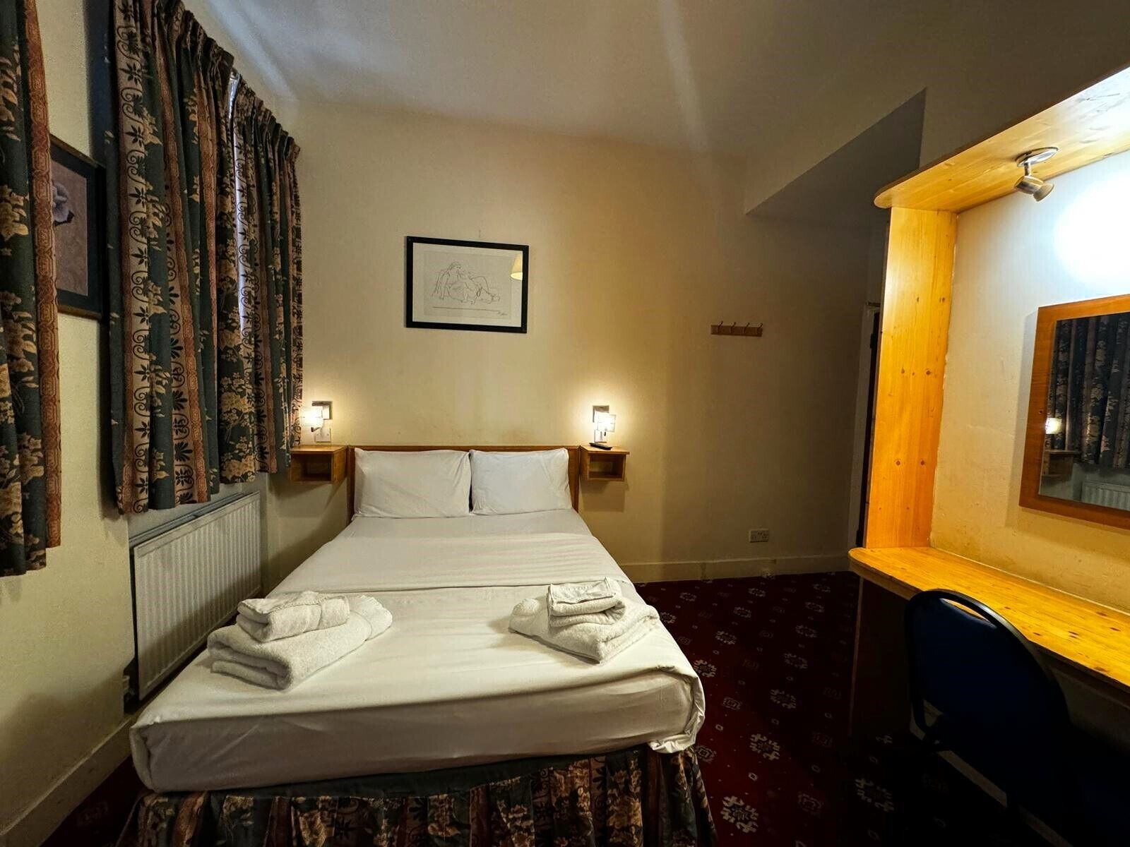 Habitación de hotel con cama, dos lámparas de noche, un cuadro en la pared y un escritorio de madera con silla.