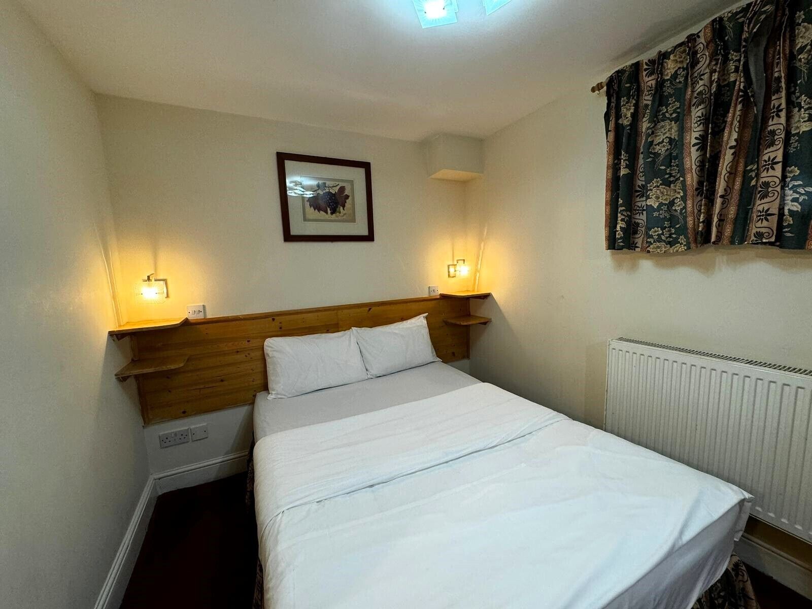 Une chambre à coucher avec une tête de lit en bois, deux lampes de chevet, un tableau encadré au mur et un radiateur.