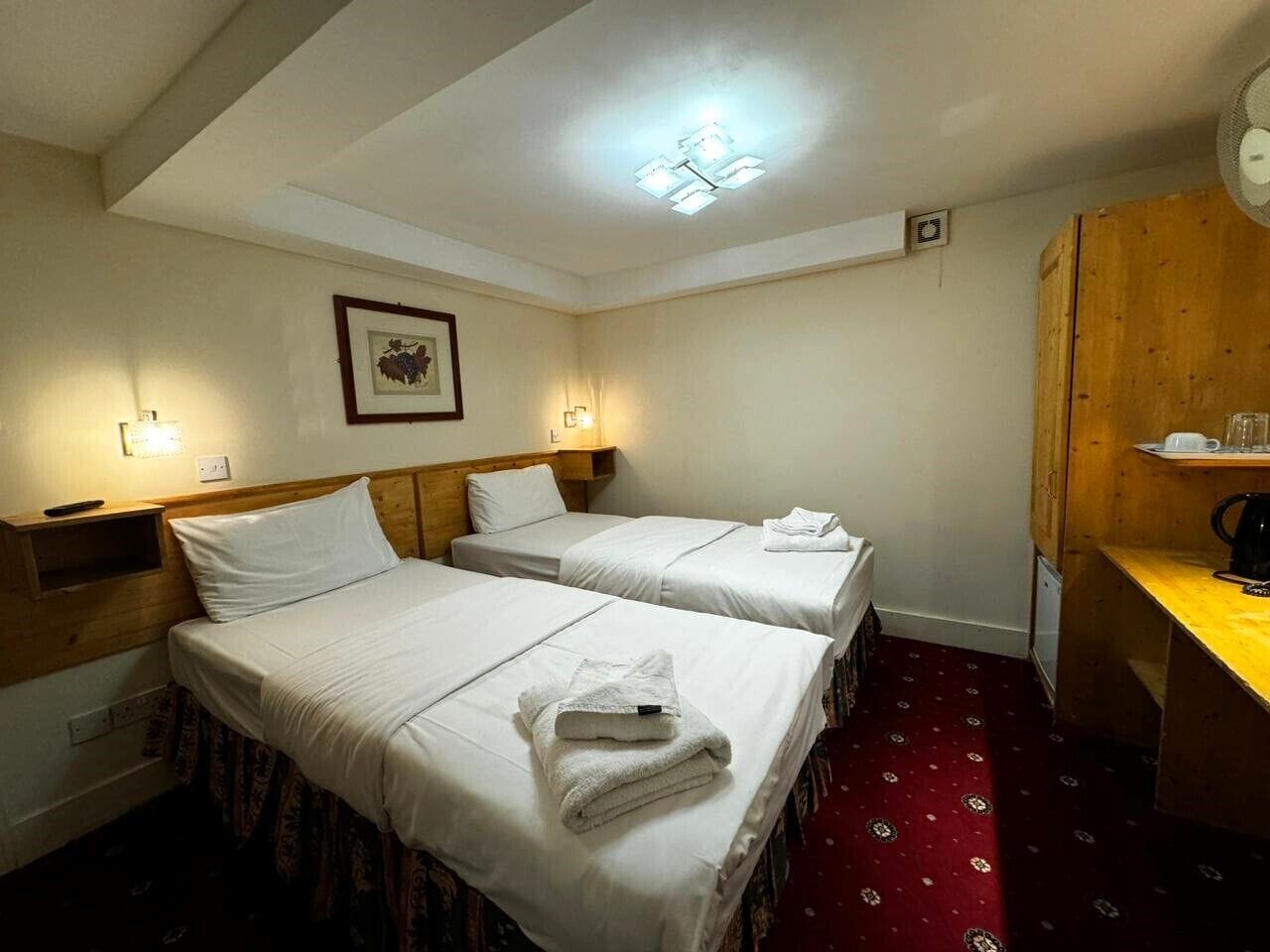 A hotel room with two beds, a wooden desk, and a red patterned carpet.