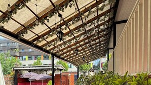 An outdoor patio with wooden tables and benches, a covered area with hanging plants, and a view of a building in the background. - Bridge Park Hotel (London)