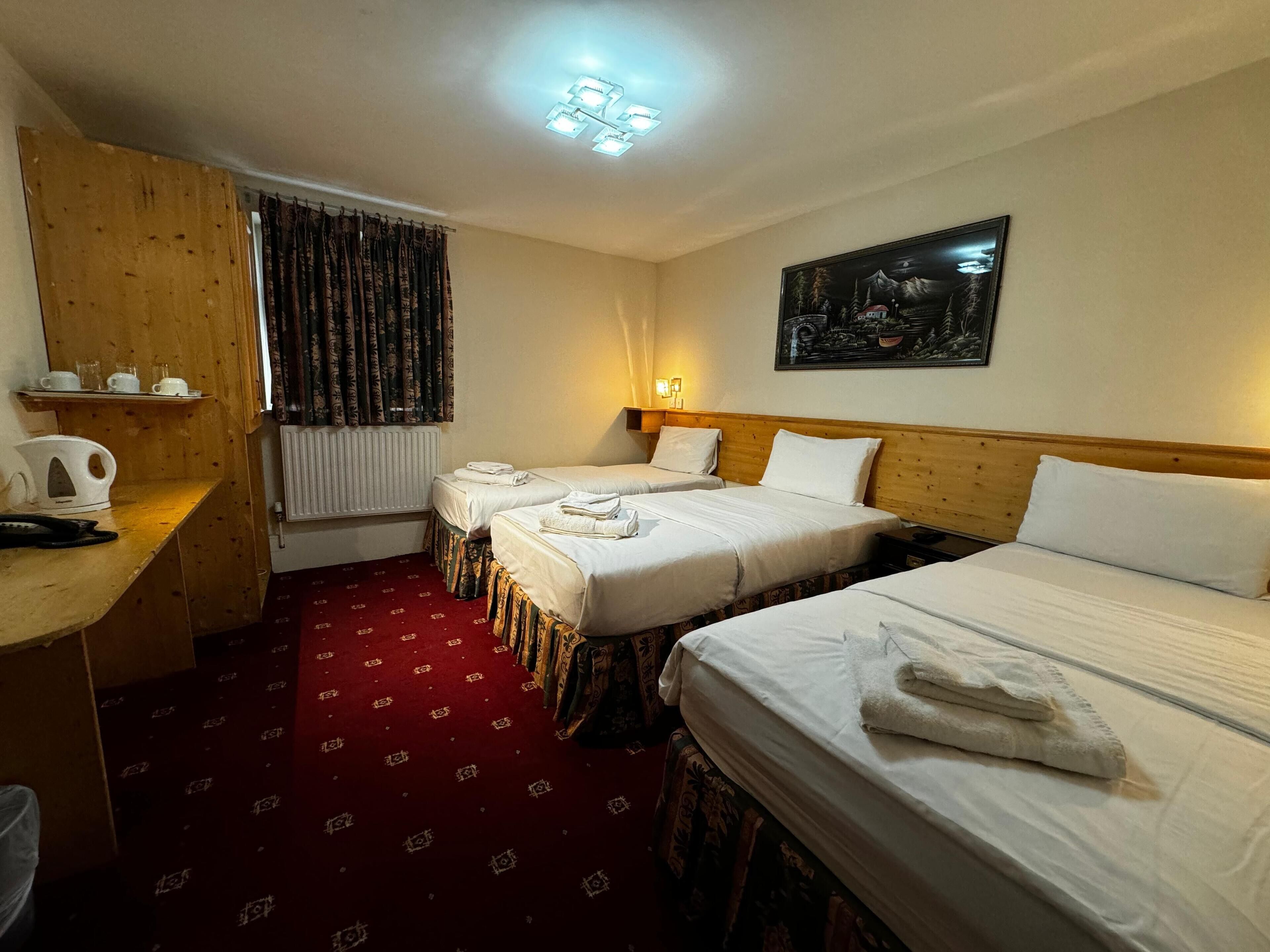 Una camera d'albergo con due letti singoli, una scrivania in legno con un bollitore, un termosifone e un quadro appeso al muro.