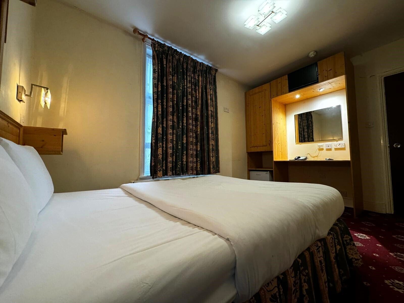 A hotel room with a large bed, a wooden wardrobe, a small desk with a mirror, and a window with patterned curtains.