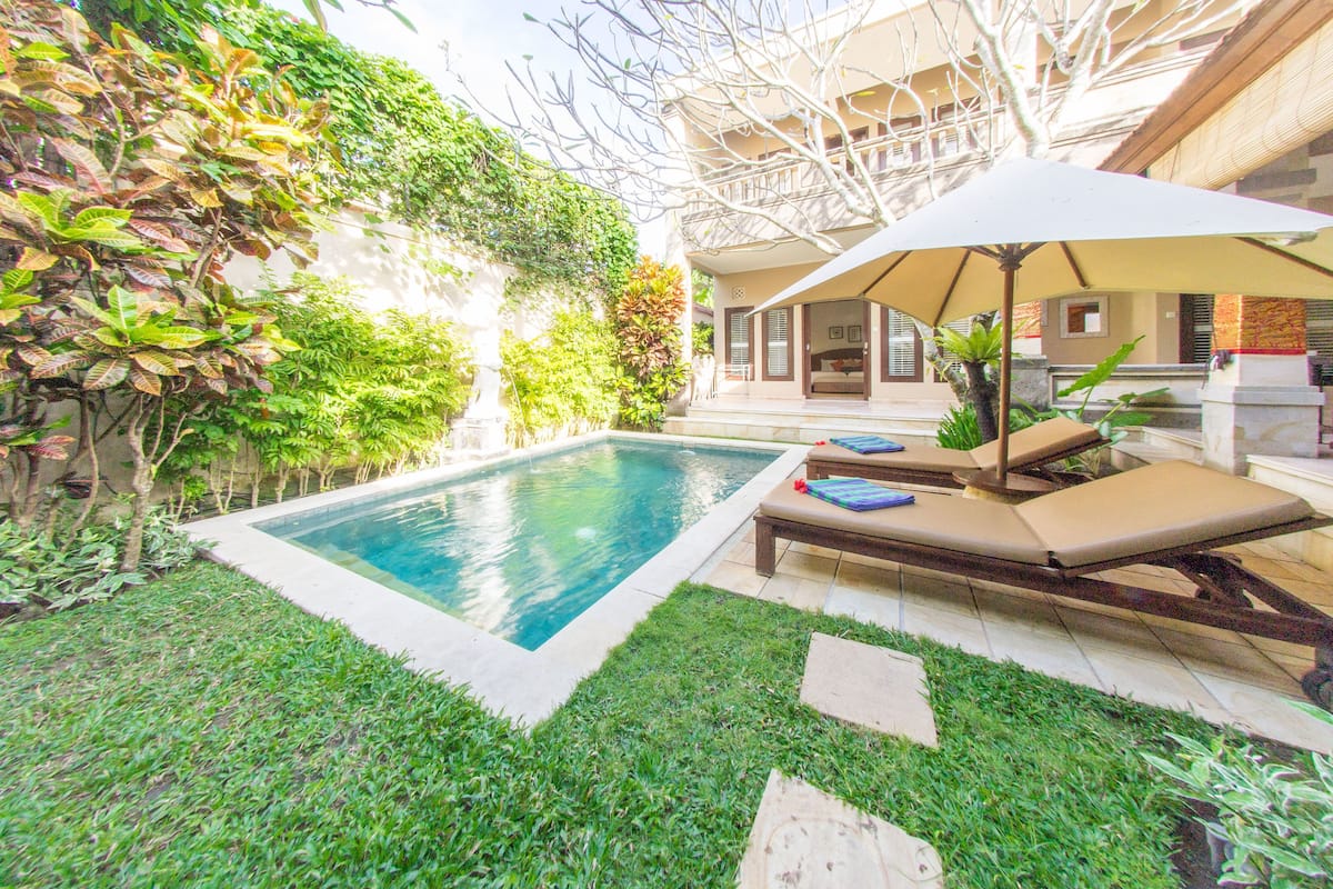 A swimming pool with lounge chairs and an umbrella, surrounded by greenery and a building with a covered patio.