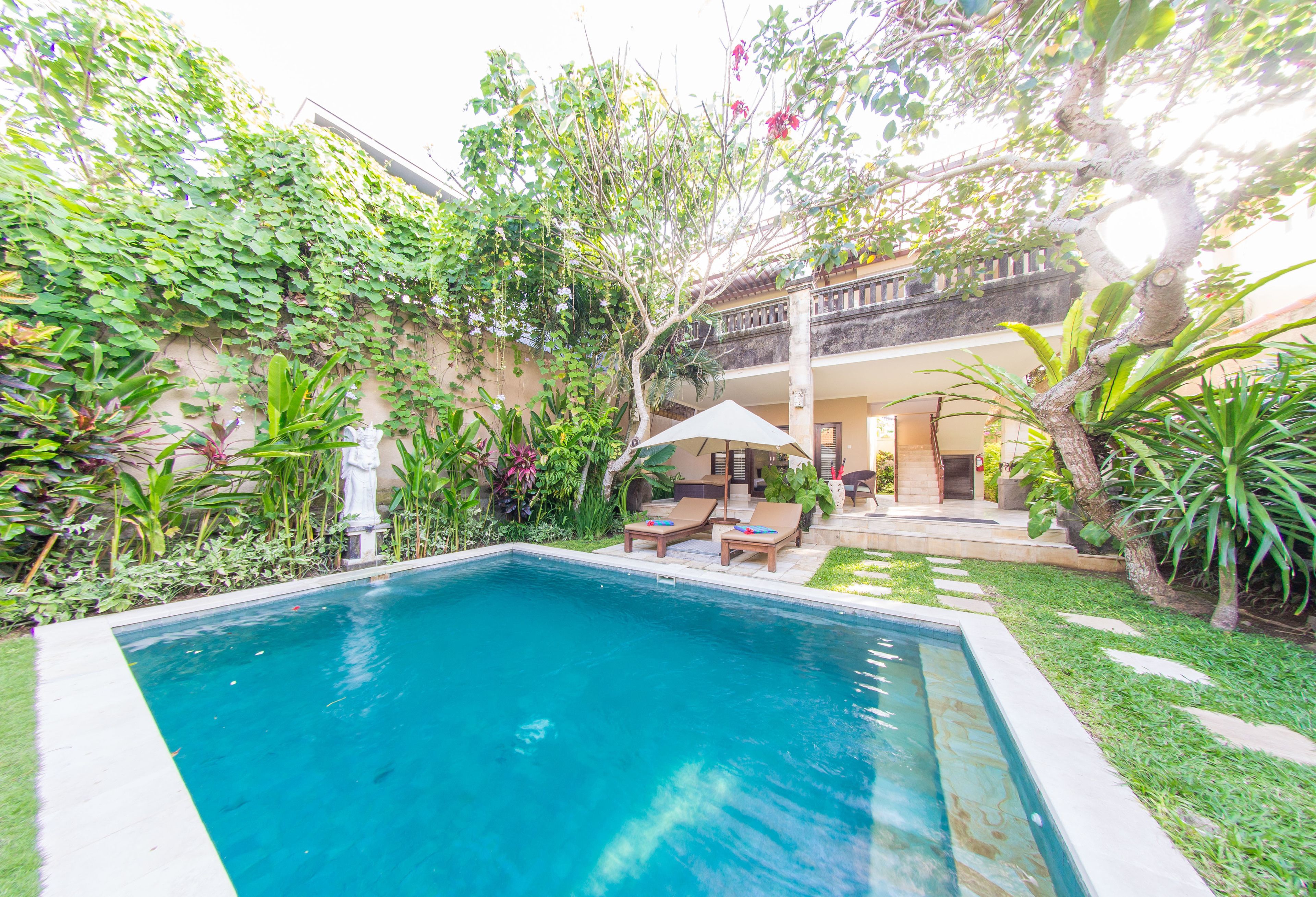A rectangular swimming pool with lounge chairs and a covered seating area, surrounded by lush greenery and a two-story building.