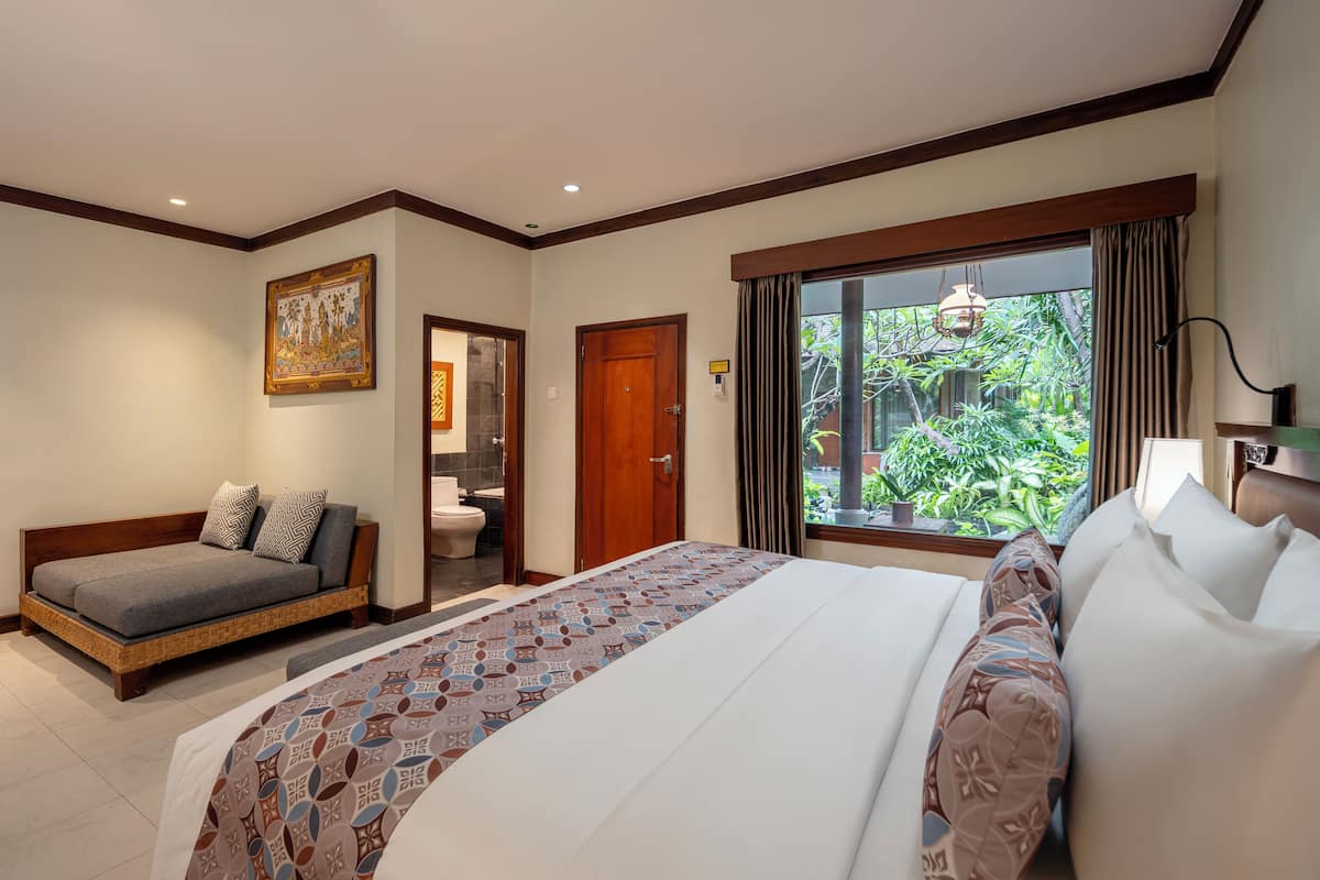 A hotel room with a large bed, a sofa, a bathroom, and a view of greenery.