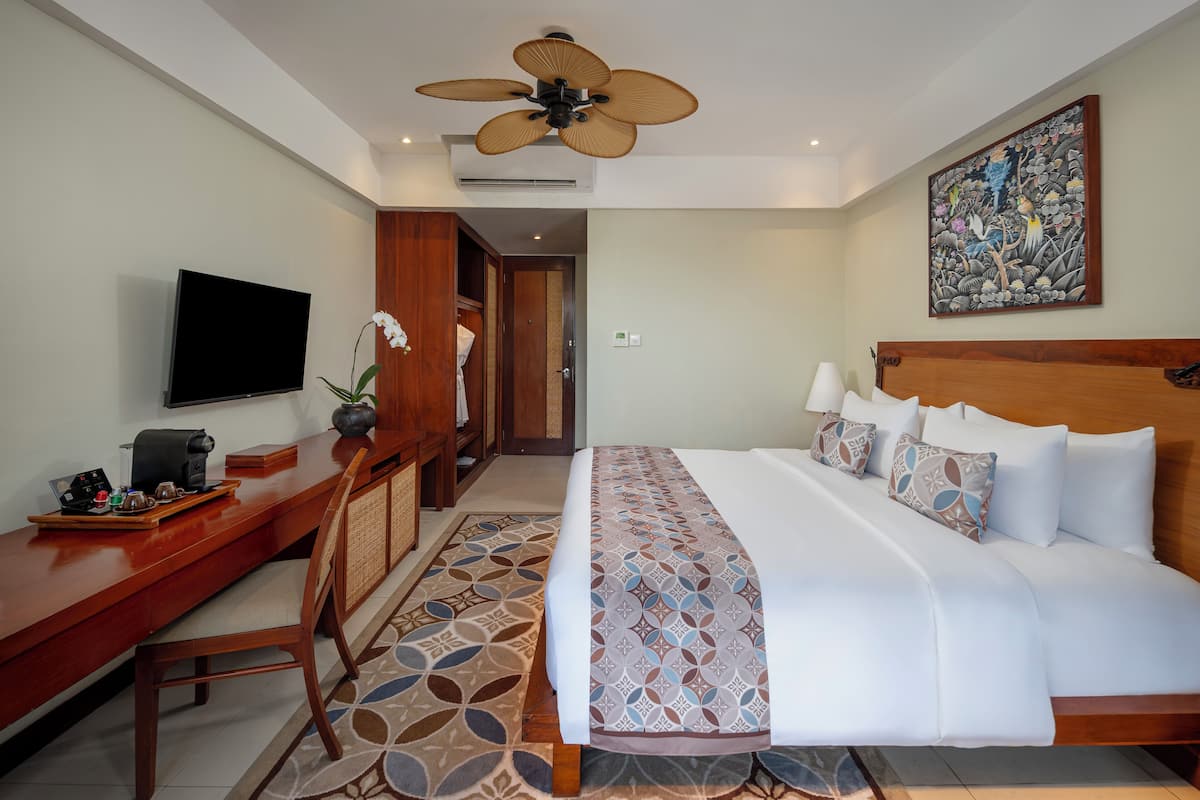 A hotel room with a bed, a desk with a TV, a ceiling fan, and a painting on the wall.
