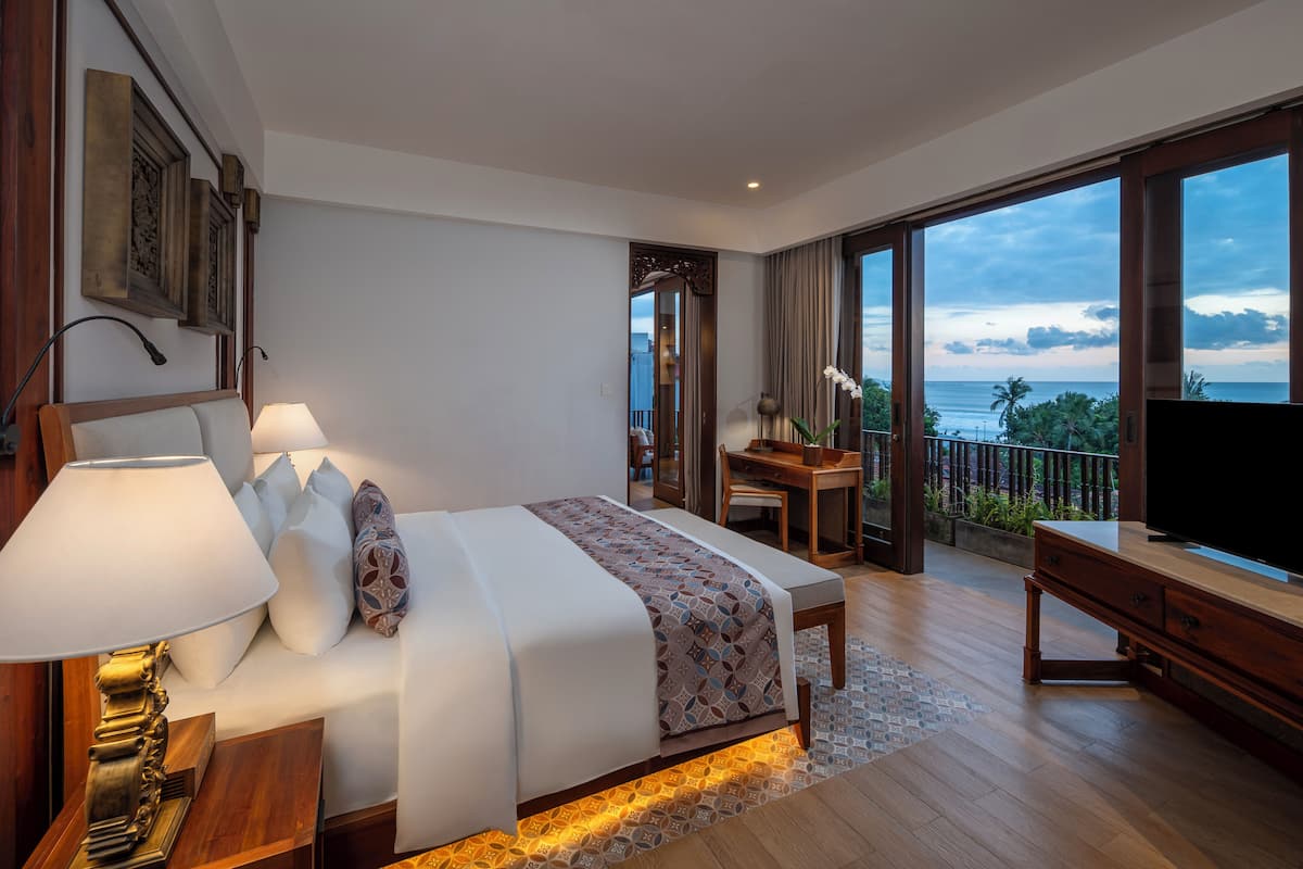 A modern hotel room with a large bed, a desk, a TV, and a balcony with a view of the ocean.