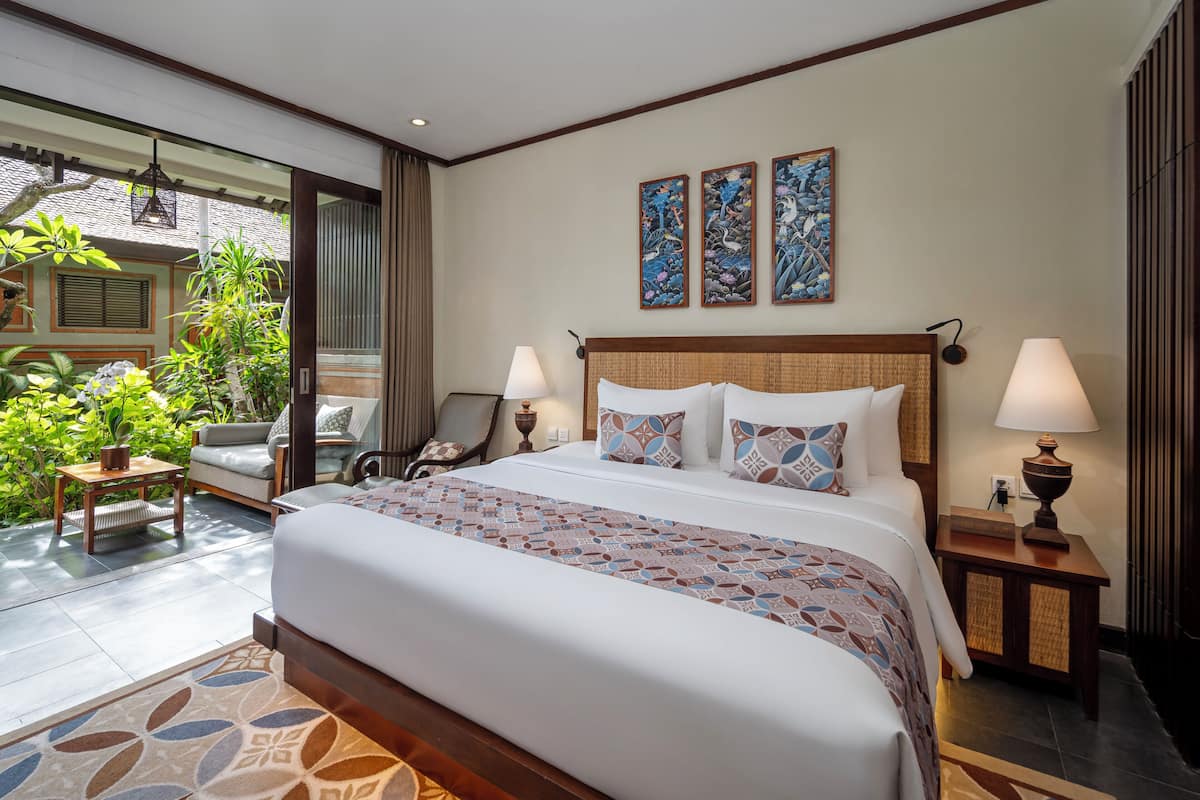 A spacious bedroom with a large bed, two bedside tables with lamps, a sofa, and a view of a lush garden.