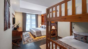 A hotel room with a bunk bed, a desk with a computer, a chair, a bed with a patterned bedspread, a bedside table with a lamp, and a window with curtains.