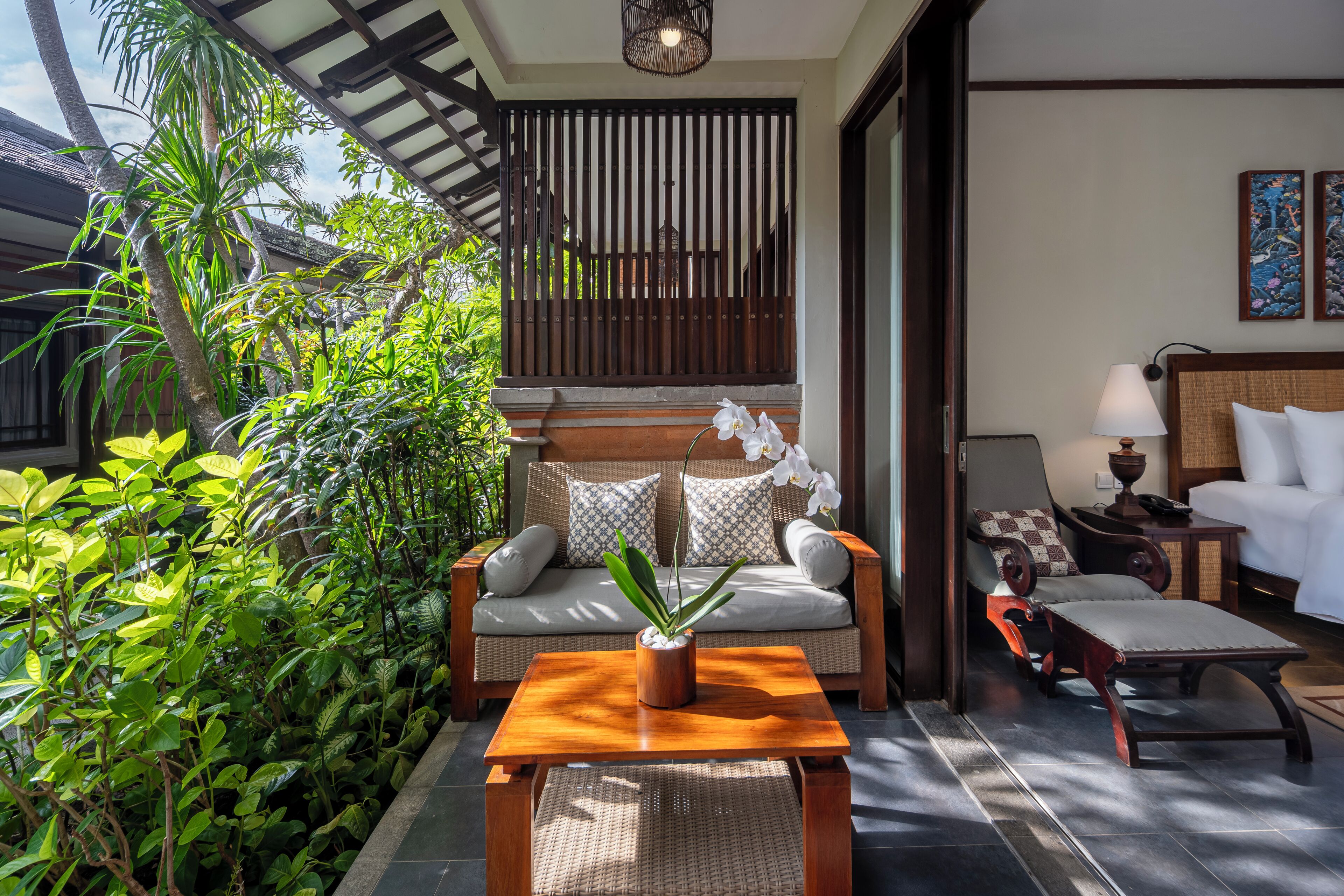 A room with a wooden sofa, a small table with a plant, a cushioned bench, and a bed with white linens.