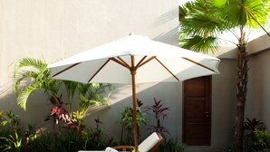 Une terrasse en bois avec deux chaises longues et un grand parasol, entourée de plantes tropicales et d’une petite piscine.