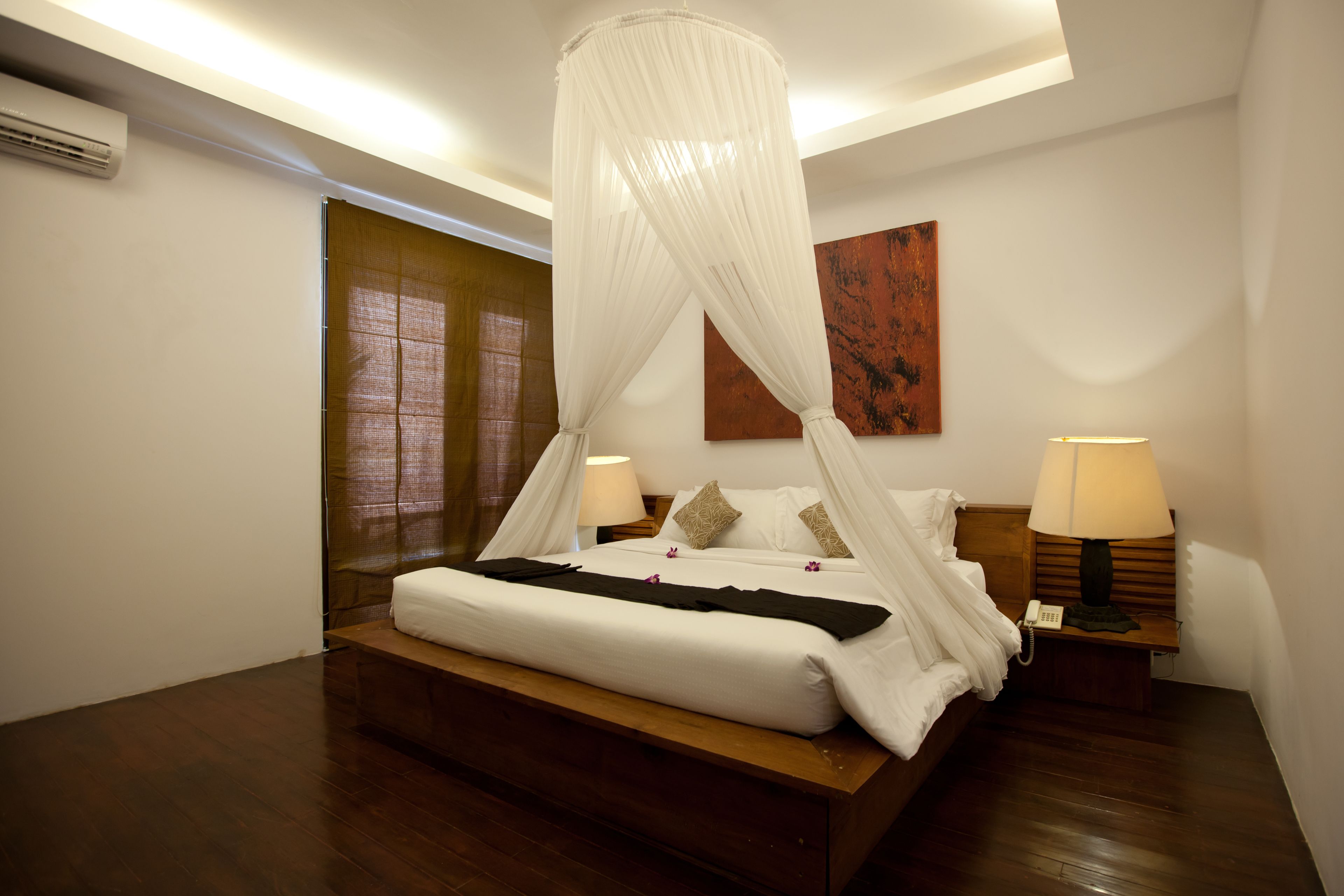 A bedroom with a four-poster bed, a wooden headboard, a bedside table with a lamp, and a wall-mounted air conditioner.