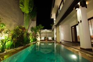 Un espace piscine nocturne avec des chaises longues et un parasol, entouré d’une végétation luxuriante et de la façade d’un bâtiment.