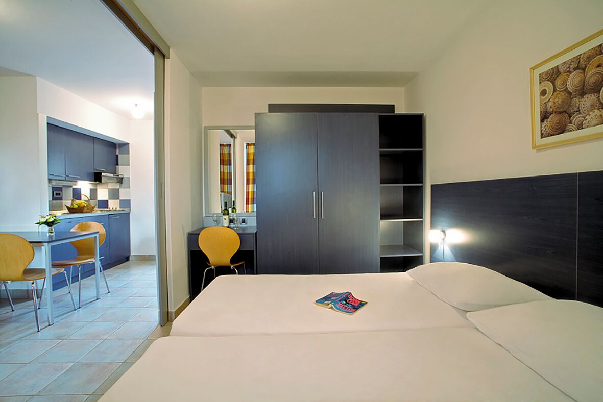 A hotel room with a bed, a desk with a chair, a wardrobe, and a kitchen area.