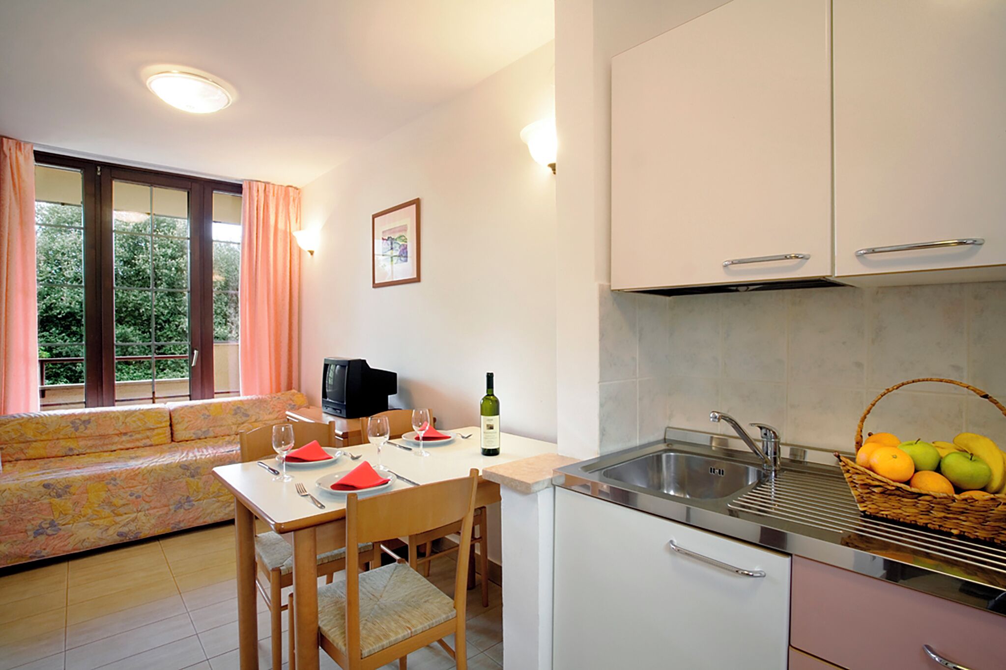 Una cocina compacta con comedor, una ventana con cortinas y un lavabo con cesta de frutas.