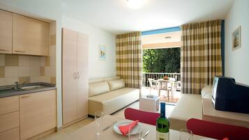 A compact hotel room with a kitchenette, a dining area, and a balcony with a view of greenery.